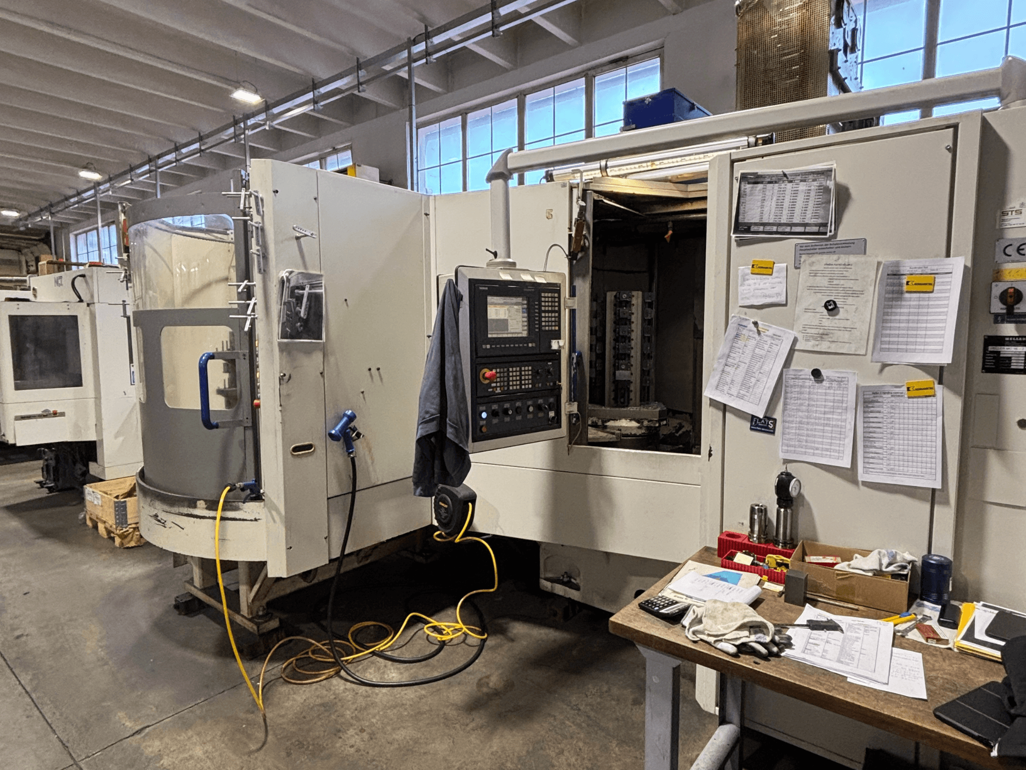 Máquina CNC en un taller, vista frontal, con panel de control y documentación en el lateral; herramientas y materiales sobre la mesa.