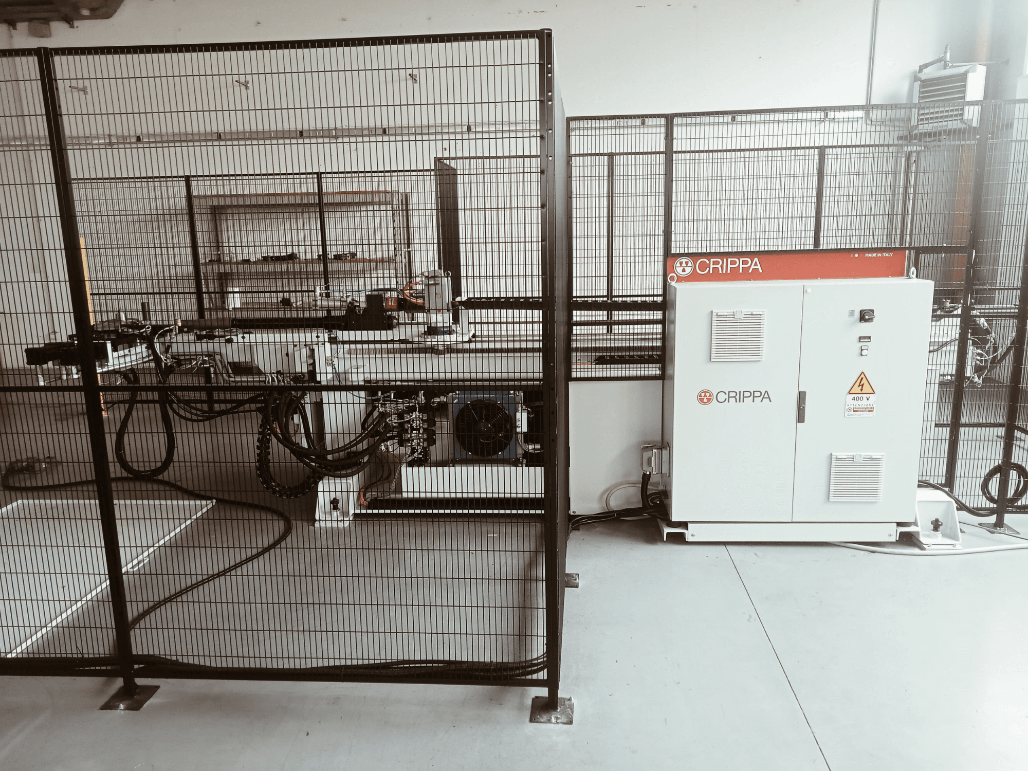 Máquina CRIPPA blanca detrás de una jaula de seguridad, con mangueras y componentes visibles, vista lateral, entorno industrial.