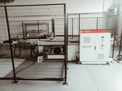 Máquina CRIPPA blanca detrás de una jaula de seguridad, con mangueras y componentes visibles, vista lateral, entorno industrial.