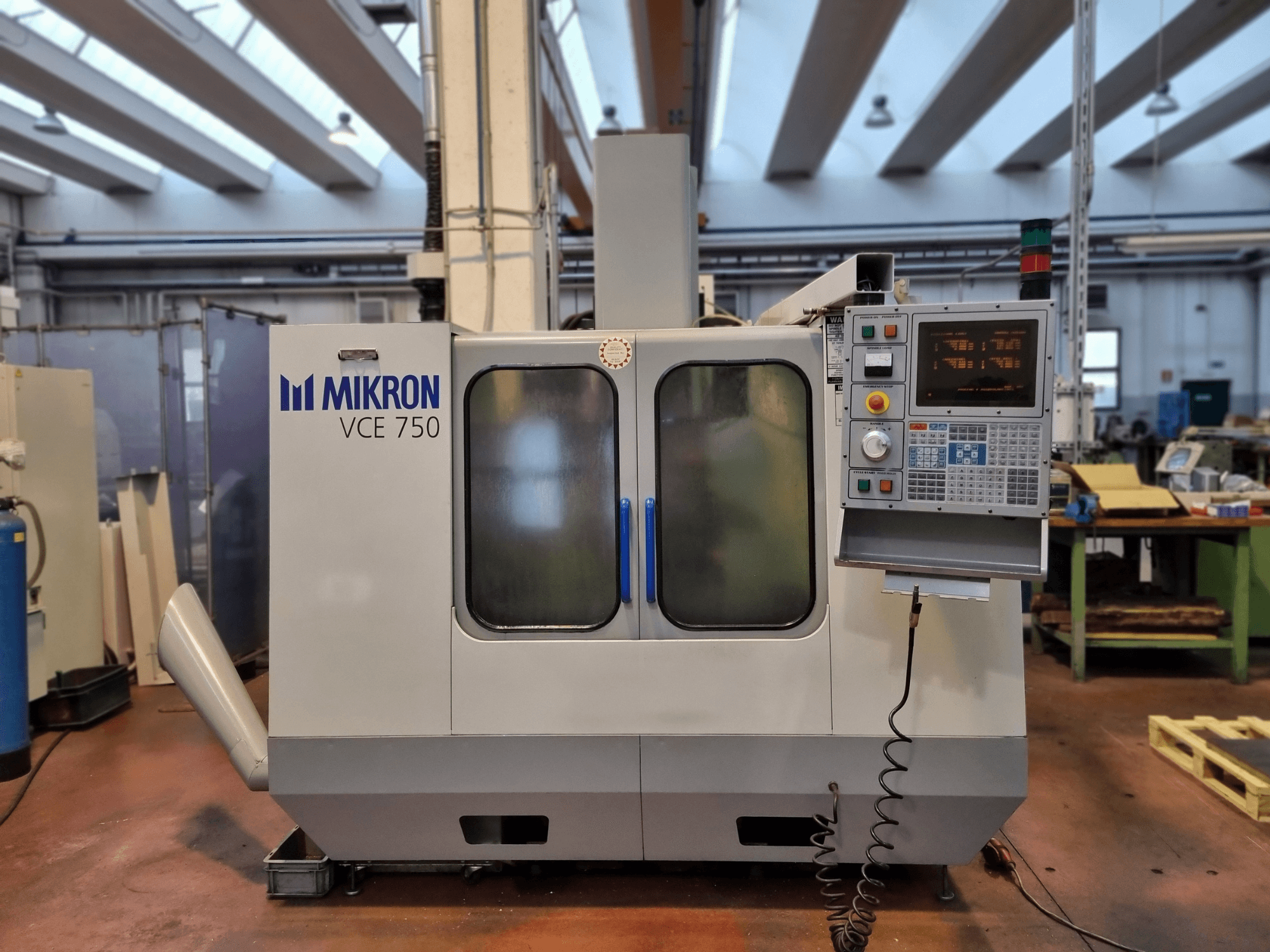 White Mikron VCE 750 CNC machine in a workshop, side view, with a digital control panel and large glass doors.