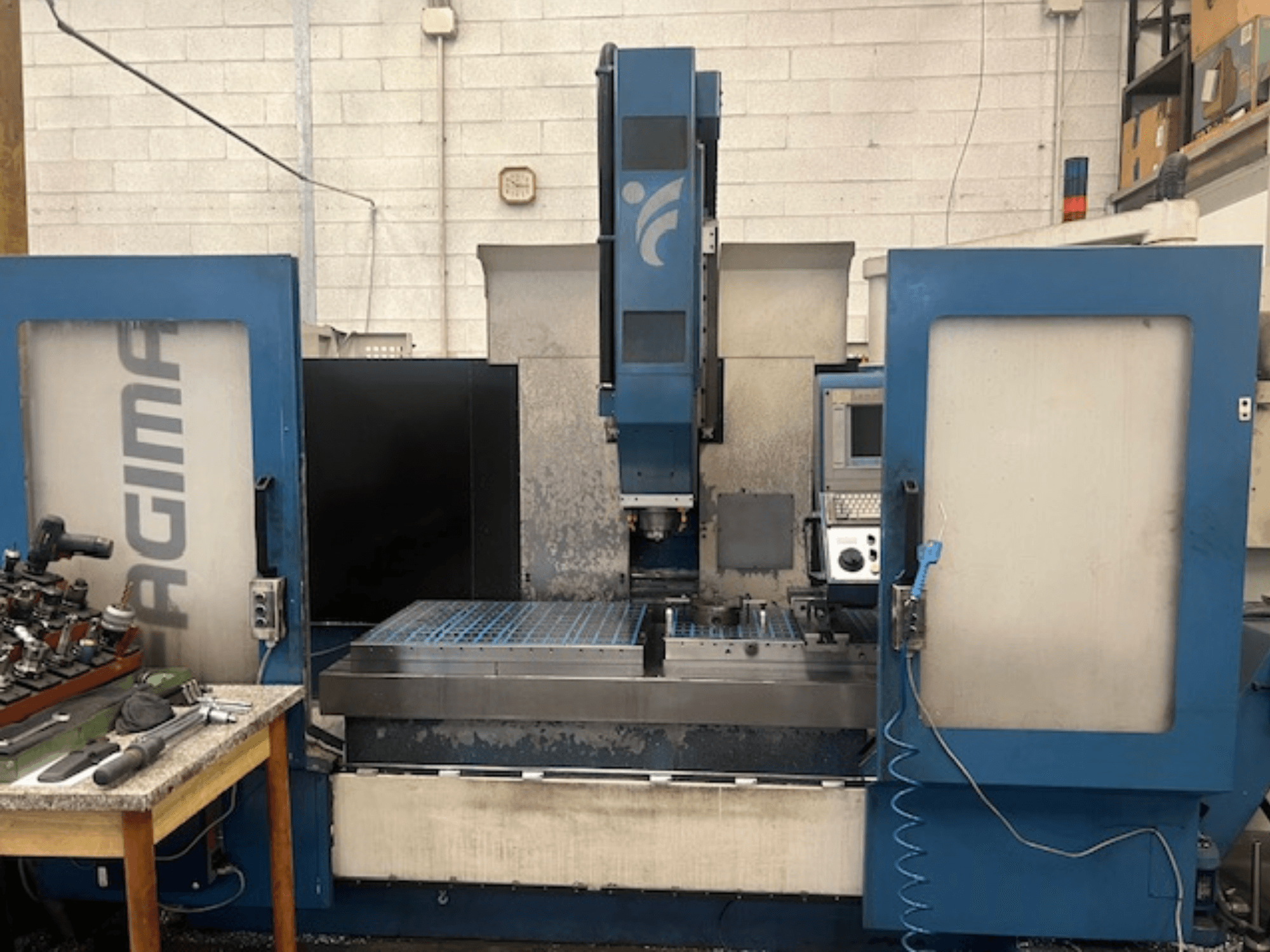 Vista frontal de una máquina CNC JAGIMA con un gran espacio de trabajo metálico, un panel de control visible y herramientas en una mesa cercana.