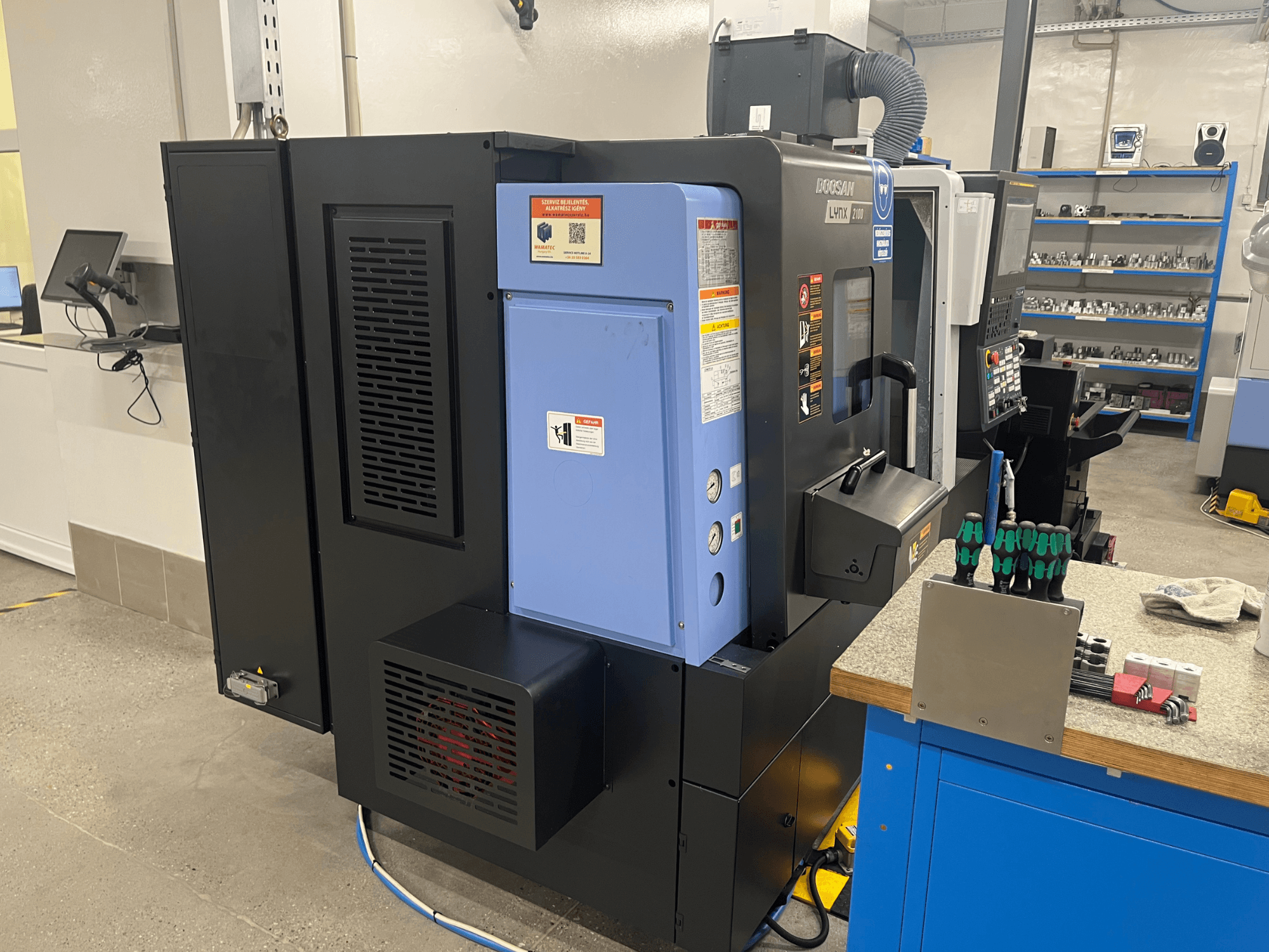 Torno Doosan Lynx 2100, vista lateral, mostrando el panel de control, el sistema de ventilación y las herramientas en un banco de trabajo.