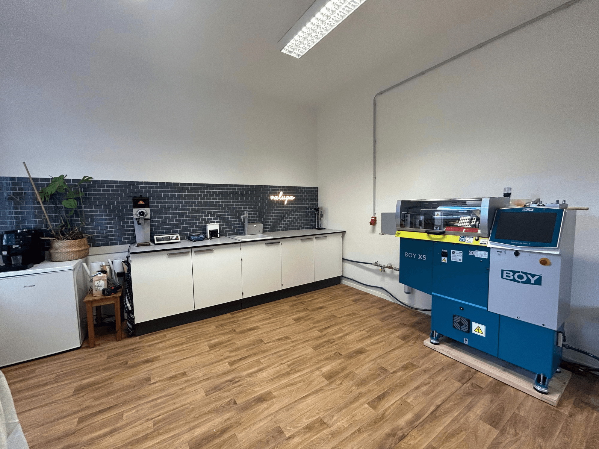 Máquina de moldeo por inyección hidráulica BOY XS 161, vista frontal, en un entorno de cocina moderna con electrodomésticos y suelo de madera.