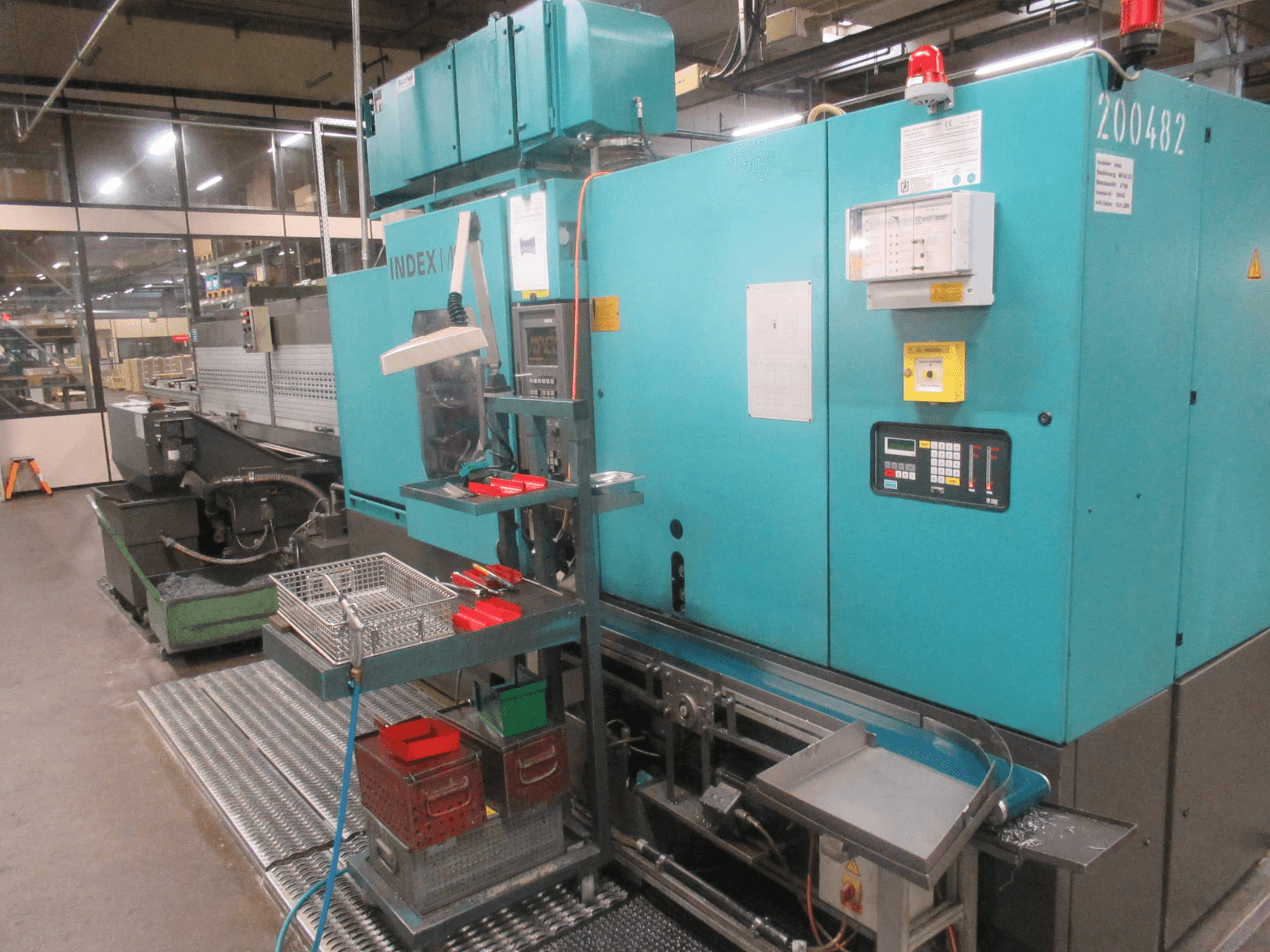 Máquina INDEX turquesa en fábrica, vista lateral; incluye panel de control, almacenamiento de herramientas y cinta transportadora. Cubos, mangueras y alarmas visibles.