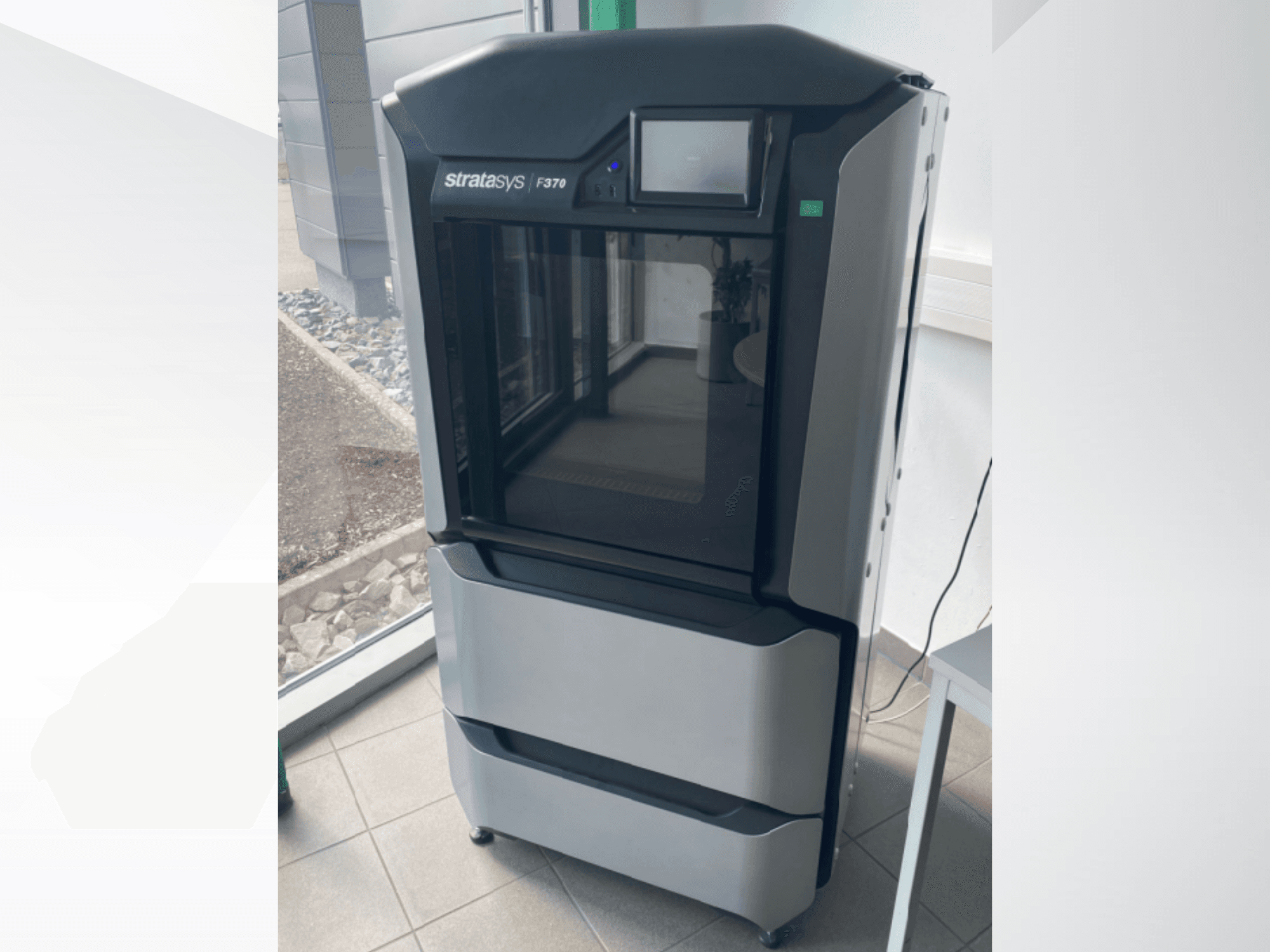 Vista frontal de la impresora 3D de plástico Stratasys F370, con un elegante diseño en negro y gris y una pantalla táctil.