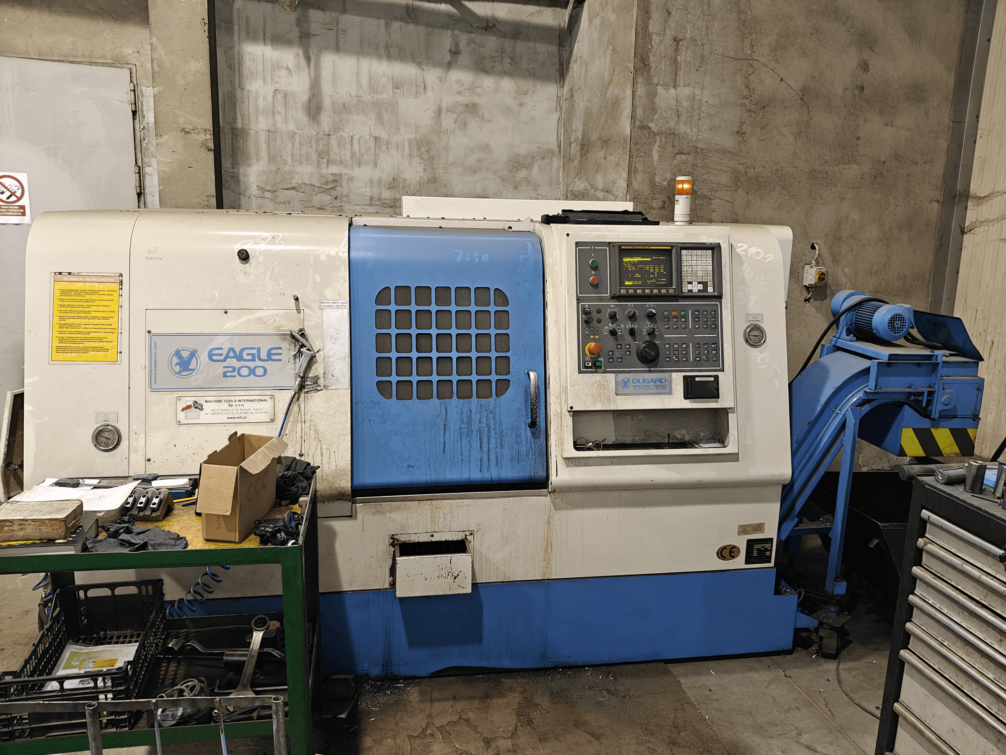 Máquina CNC Eagle 200, vista frontal con panel de control, varias herramientas en un carro verde. Desgaste visible y luces de funcionamiento.