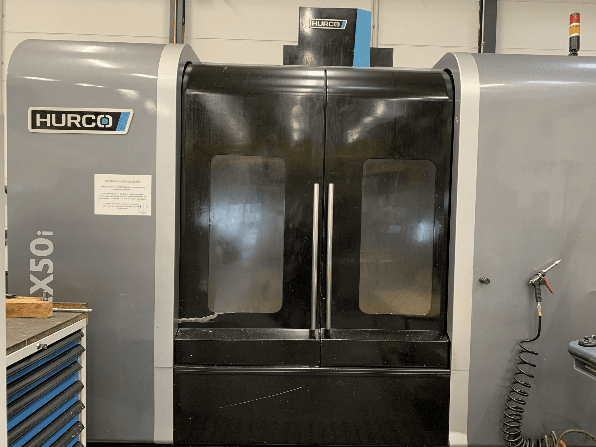 Vista frontal de una máquina CNC Hurco VX50i con puertas dobles, que muestra los controles técnicos y un letrero de mantenimiento en el lateral.