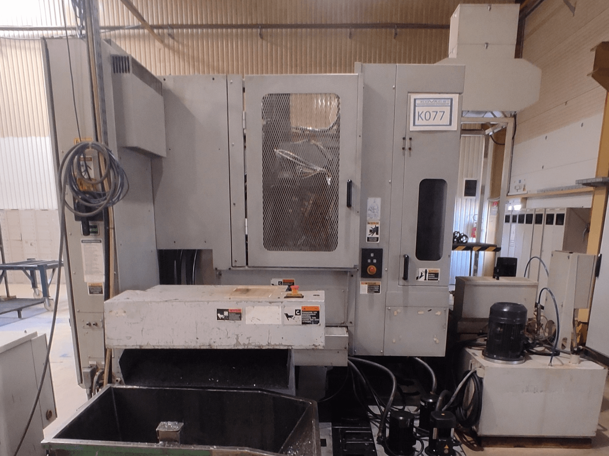 Gray industrial CNC machine with a large worktable, side view, featuring control panel and coolant reservoir, model K077.