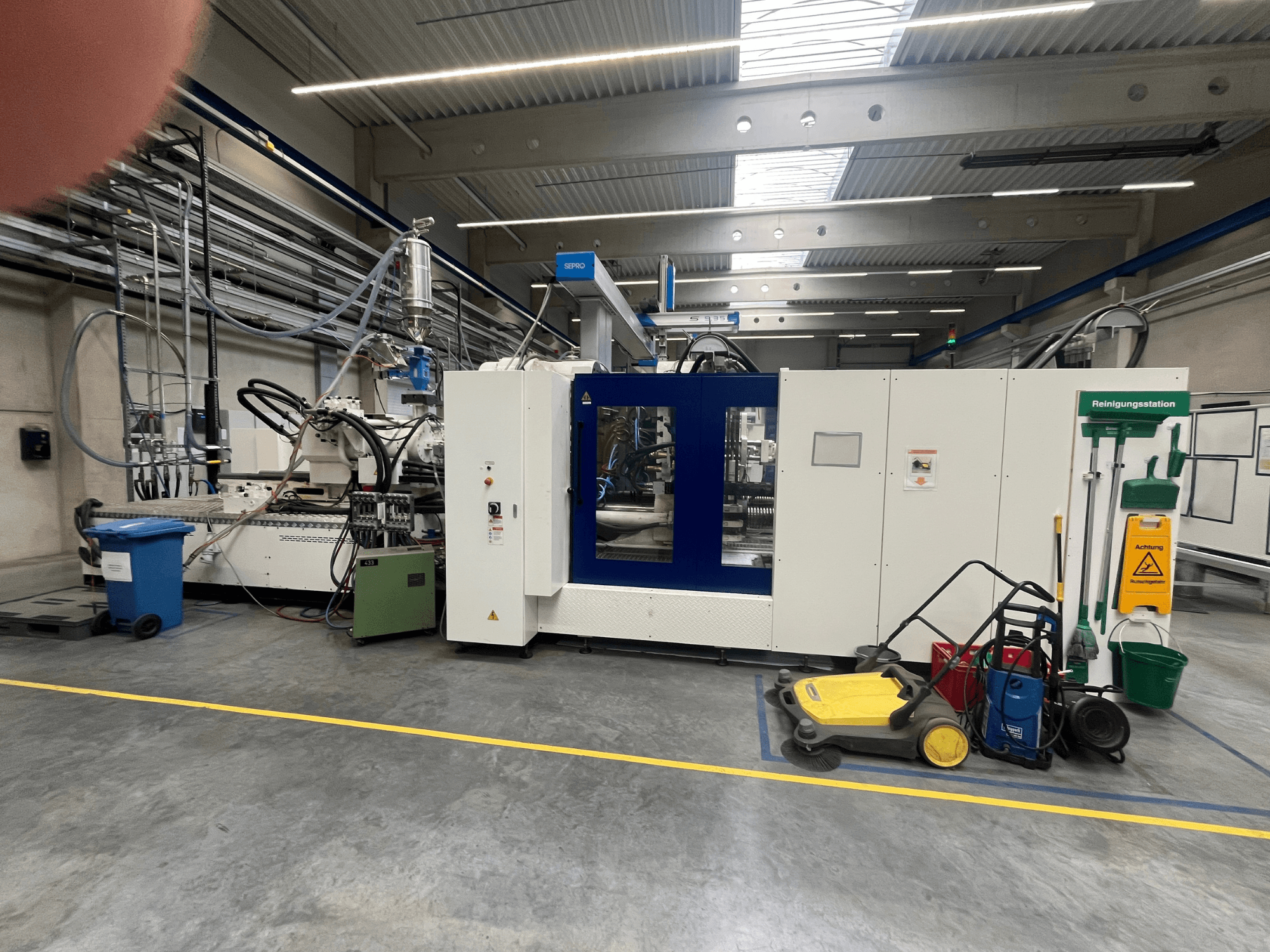 Máquina industrial en una fábrica, vista frontal, con paneles azules y blancos, mangueras y equipo de limpieza cerca.