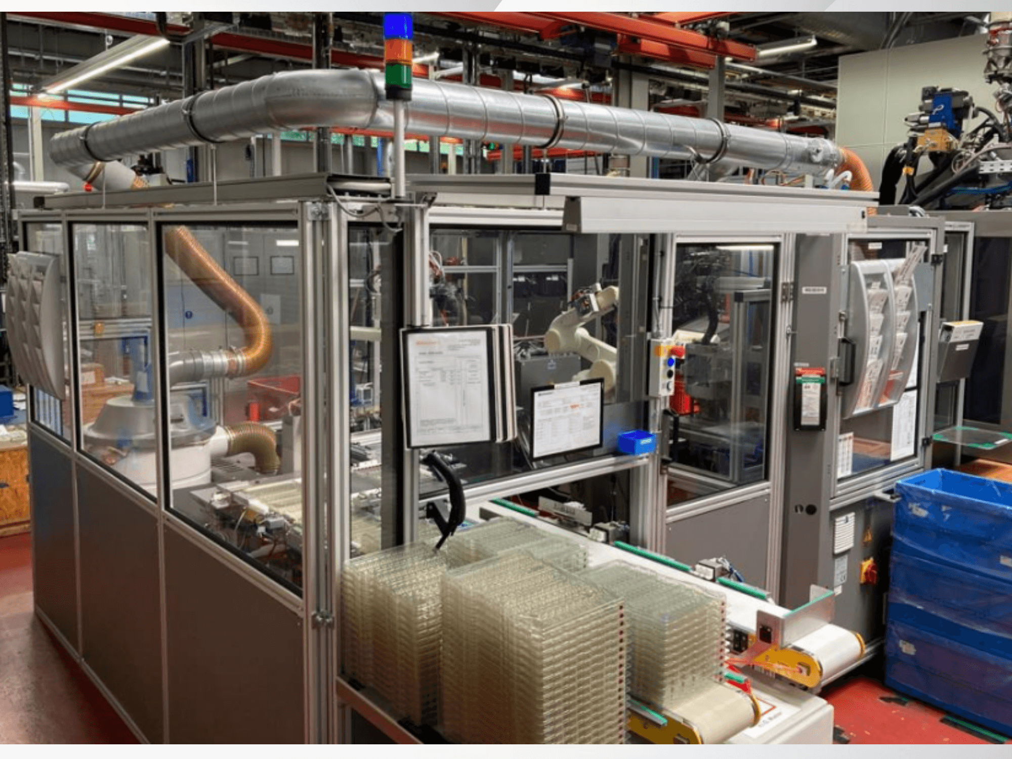 Vista frontal de una máquina automatizada con tubos metálicos, panel de control y contenedores de plástico transparente apilados en la planta de una fábrica.