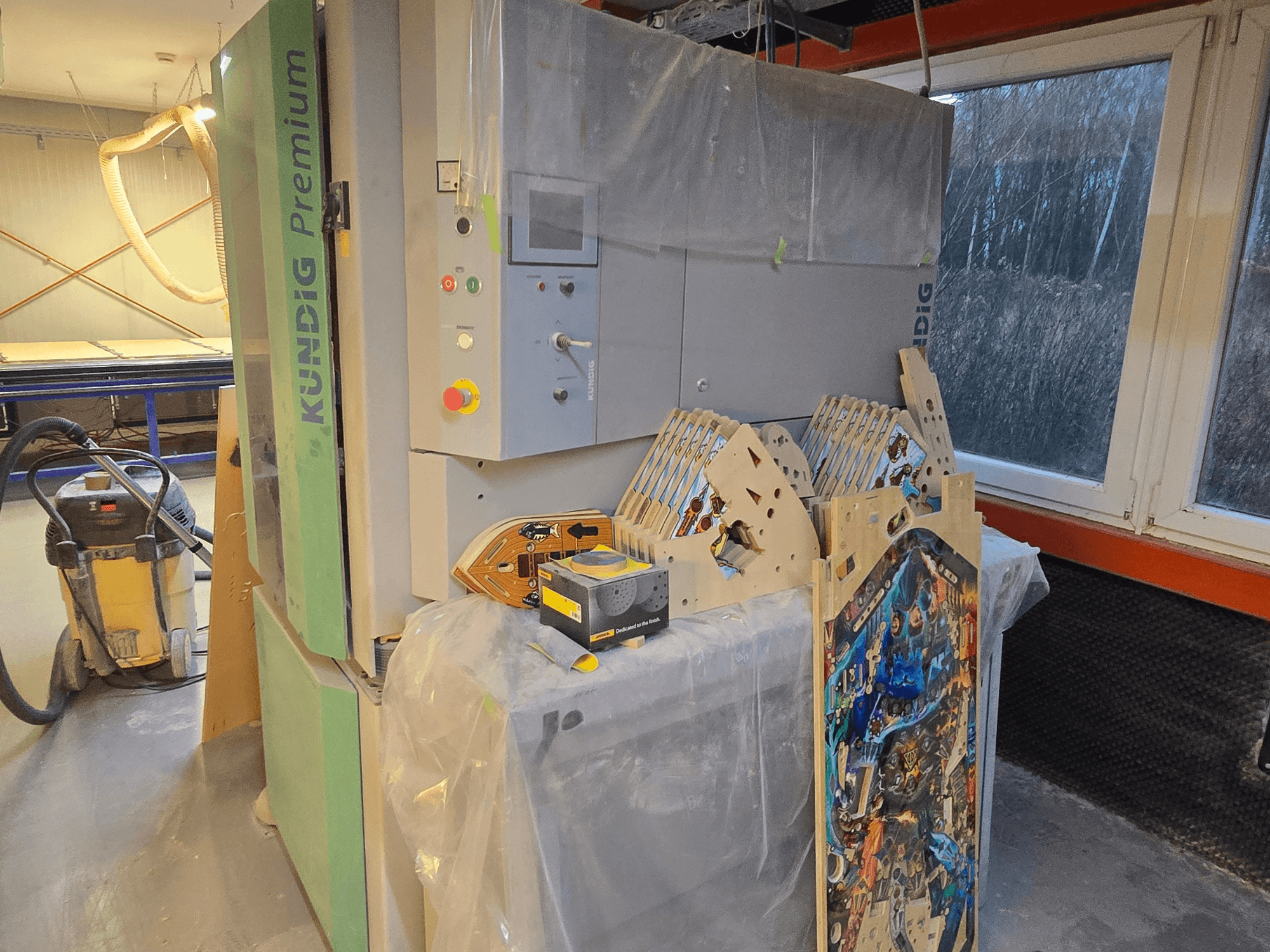 Lijadora de banda Kundig Premium-1 1100-C en el taller, mostrando el panel de control y las piezas de madera, vista lateral, con aspirador de polvo al fondo.