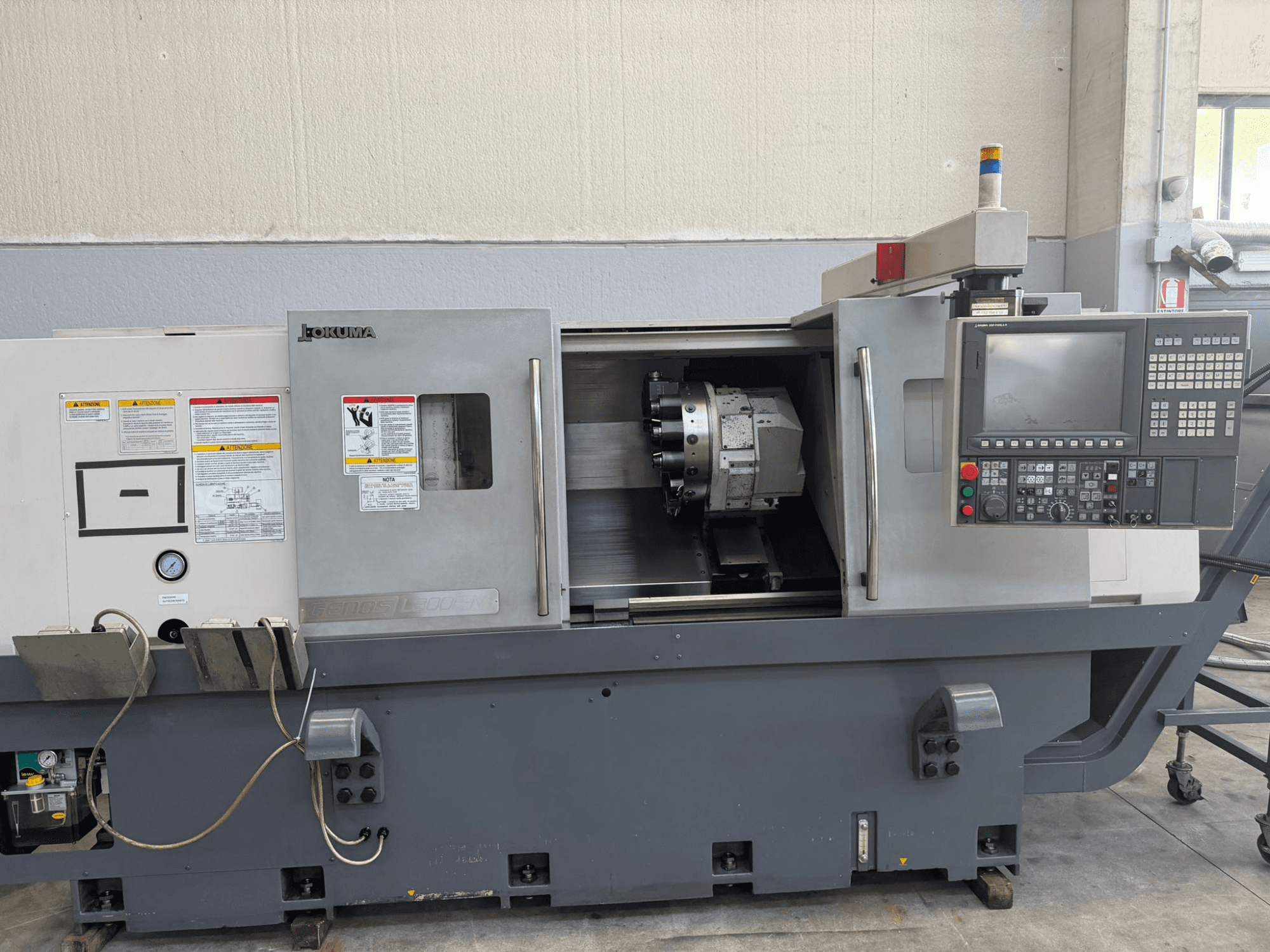 Máquina de torno CNC Okuma, vista frontal, mostrando el panel de control, el portaherramientas de corte y las advertencias de seguridad en el lateral.