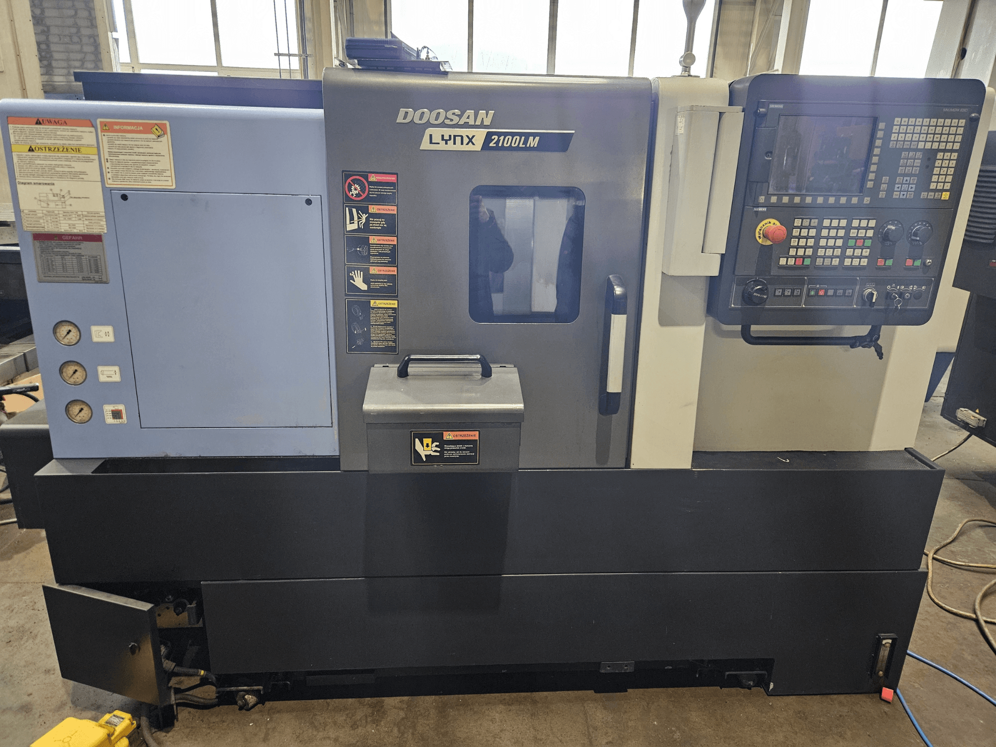 Torno CNC Doosan Lynx 2100LM, vista frontal, con panel de control, manómetros de aire y etiquetas de seguridad visibles.