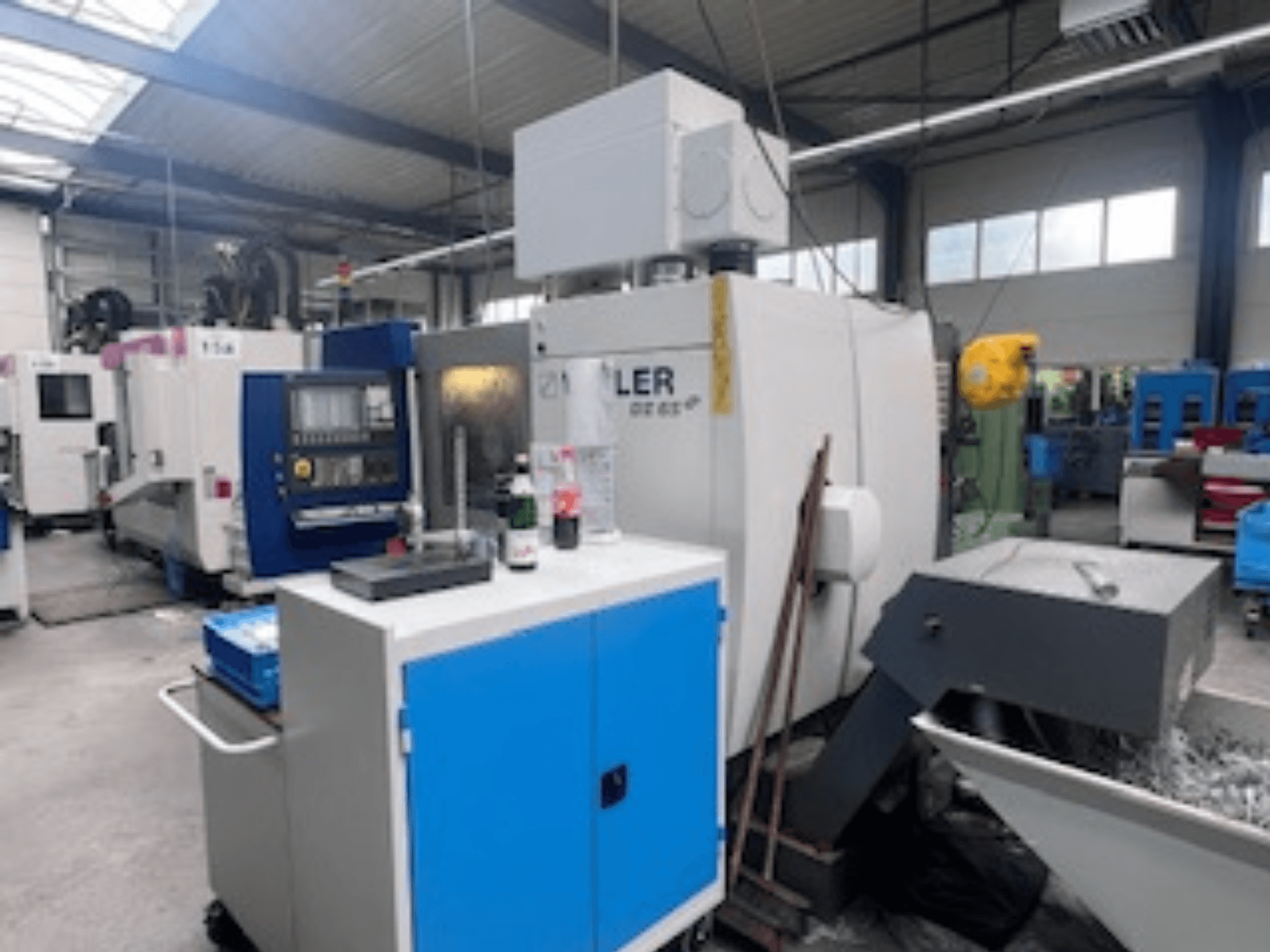 Taller industrial con un centro de mecanizado Heller, vista frontal que muestra herramientas, controles y equipos circundantes.