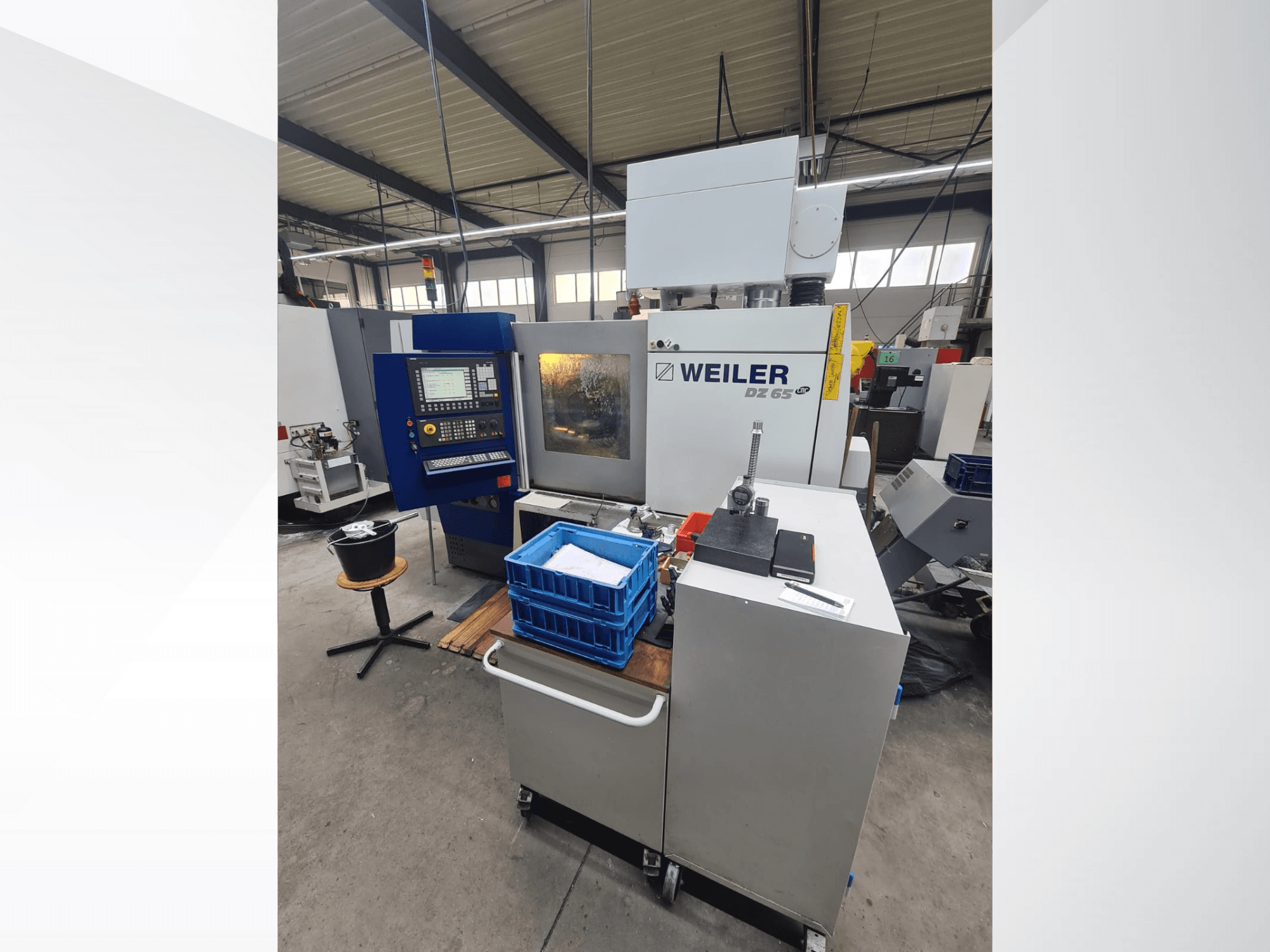 Máquina CNC Weiler DZ 65 en un taller, vista frontal con panel de control, herramientas y cubos de almacenamiento azules visibles.
