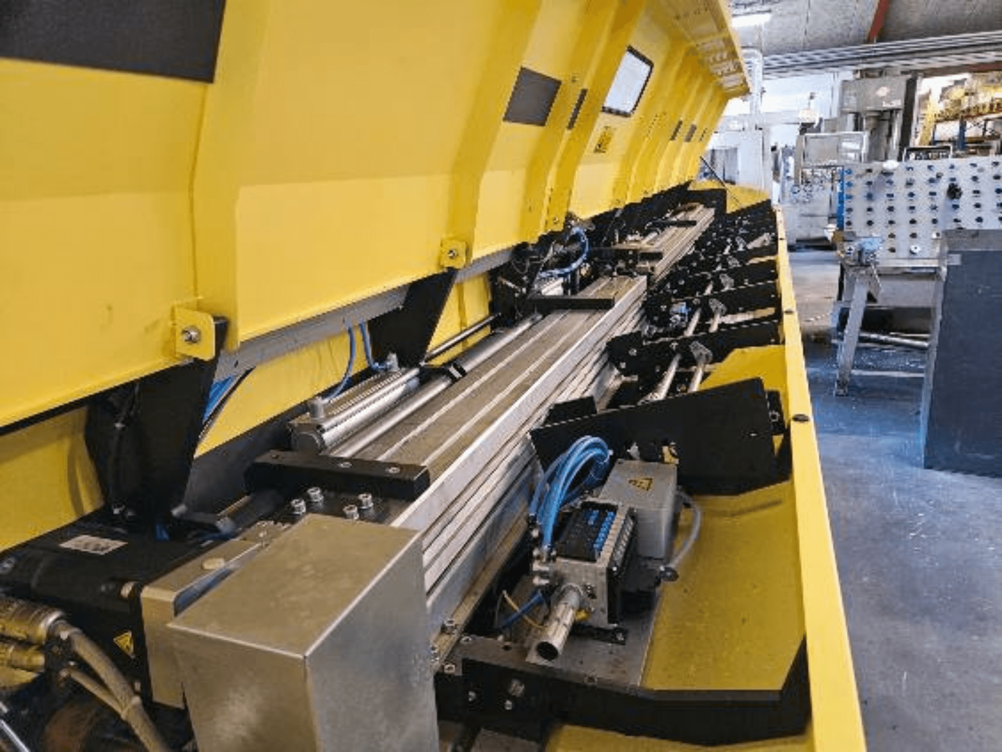 Primer plano de una máquina hidráulica amarilla con componentes metálicos y raíles transportadores, con mangueras y piezas electrónicas.