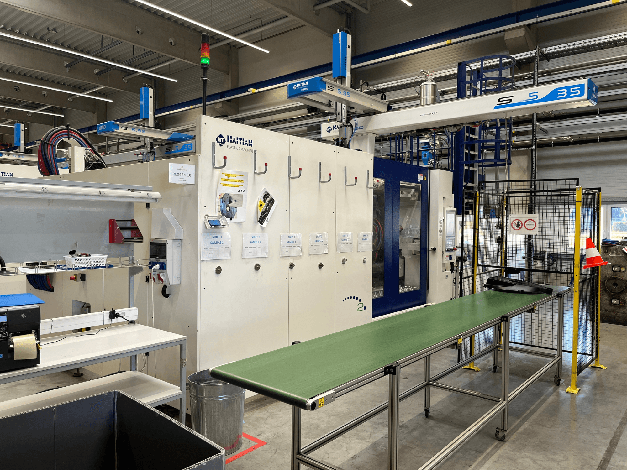 Máquina de plástico S5-35 haitiana en un taller; mesa transportadora verde, barreras de seguridad y panel de control visibles desde la vista frontal.