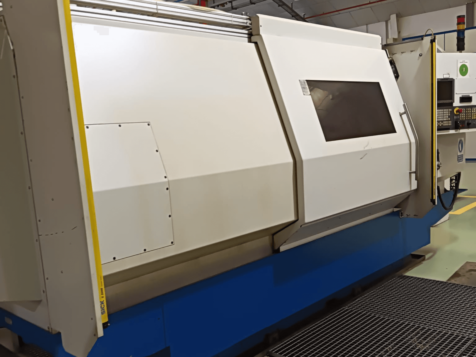 Torno industrial, vista lateral, con una gran carcasa blanca y azul, panel de control digital y dispositivos de seguridad.