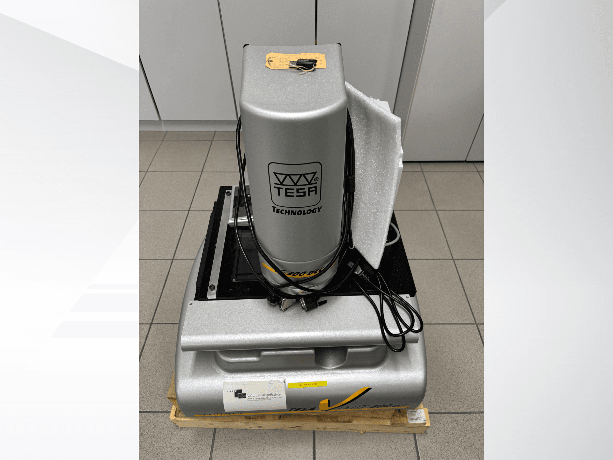 Máquina de medición óptica TESA Visio 300 DCC en una vista frontal con cables, sobre un palé de madera, y embalaje protector visible.