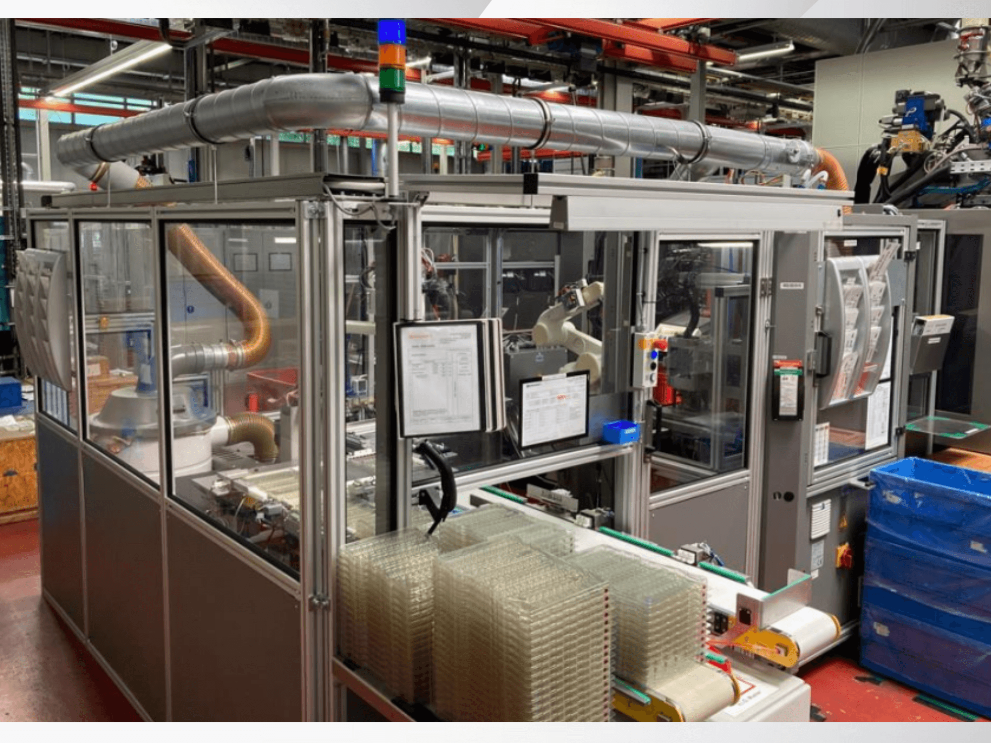Máquina de montaje automatizada con brazo robótico, paneles laterales transparentes y contenedores apilados de artículos transparentes, vista frontal.