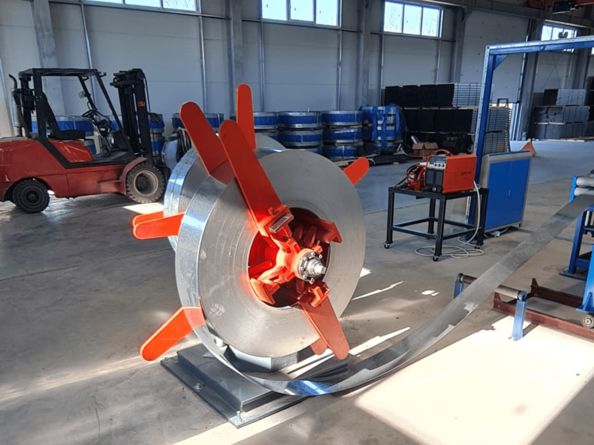 Bobina metálica con asas rojas sobre un soporte, vista desde un ángulo. Fondo industrial con maquinaria y una carretilla elevadora.