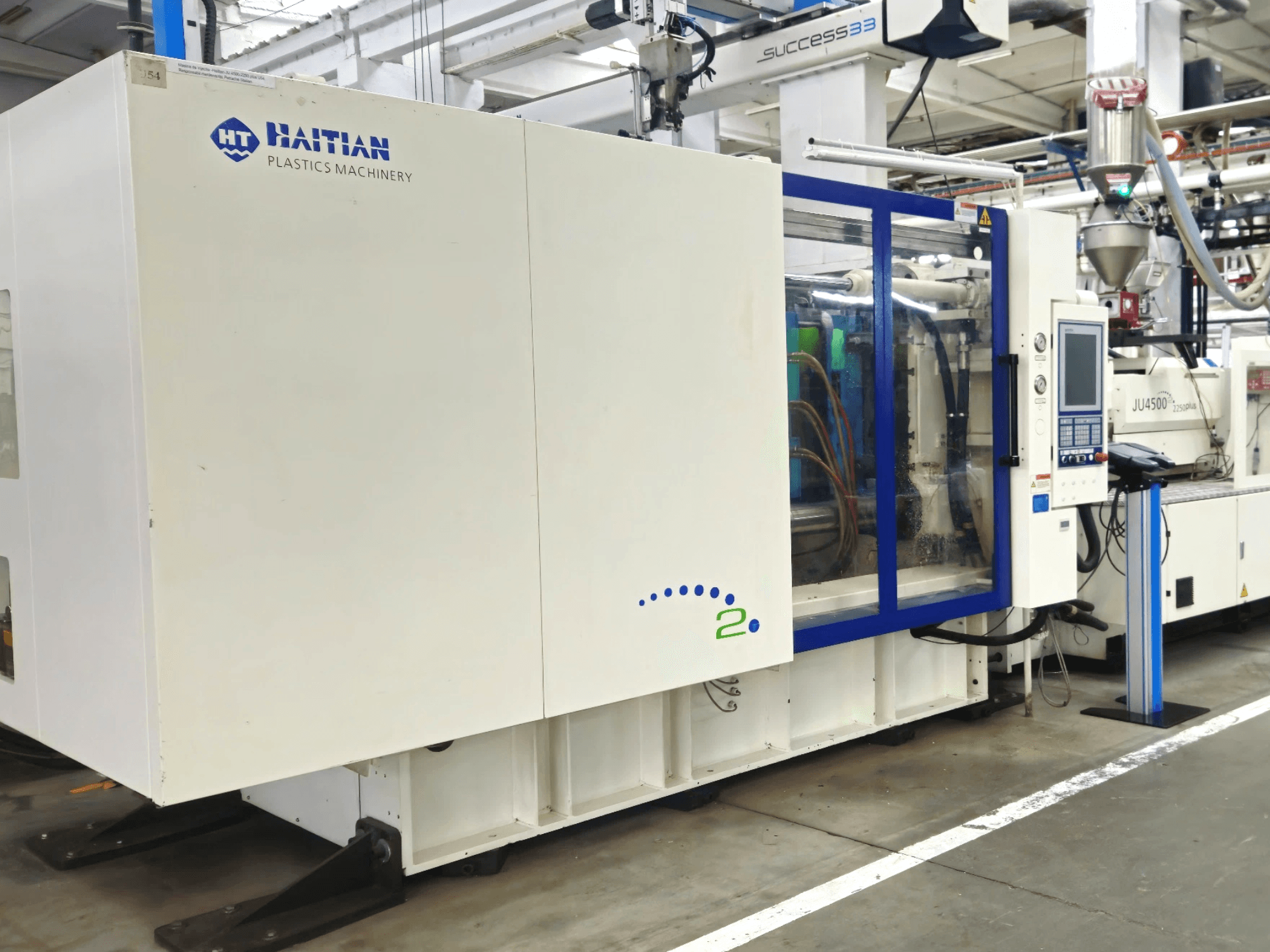 Maqueta de Haitian Plastics Machinery, vista frontal, mostrando maquinaria con panel de control y tubos visibles, en un entorno industrial.