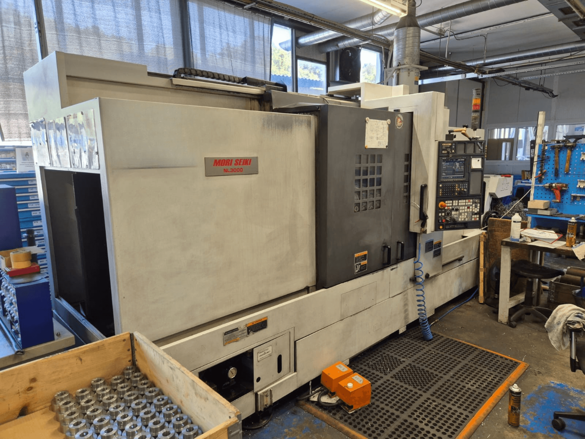 Torno CNC Mori Seiki NL3000 en un taller, vista lateral mostrando el panel de control, herramientas y piezas metálicas en una caja de madera.