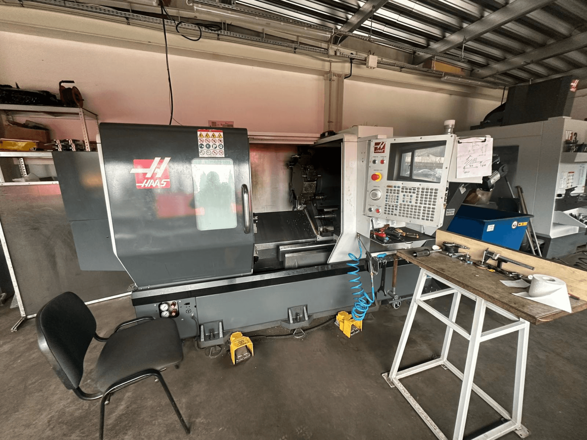 Torno CNC Haas (modelo ST-10) en un taller, vista lateral mostrando controles, herramientas en un banco de trabajo y una silla cerca.