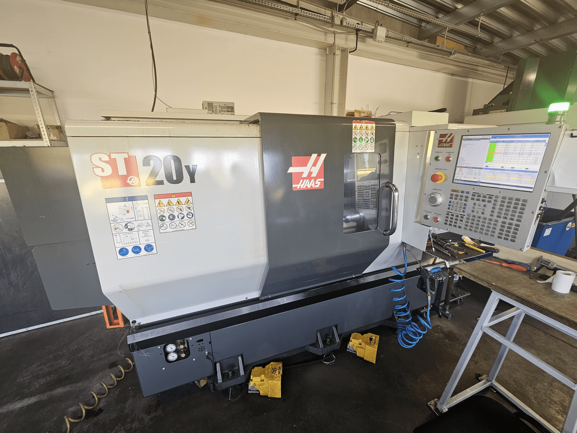 Torno CNC Haas ST-20Y en vista frontal con panel de control; incluye etiquetas de seguridad y detalle de manguera de aire en el lateral.