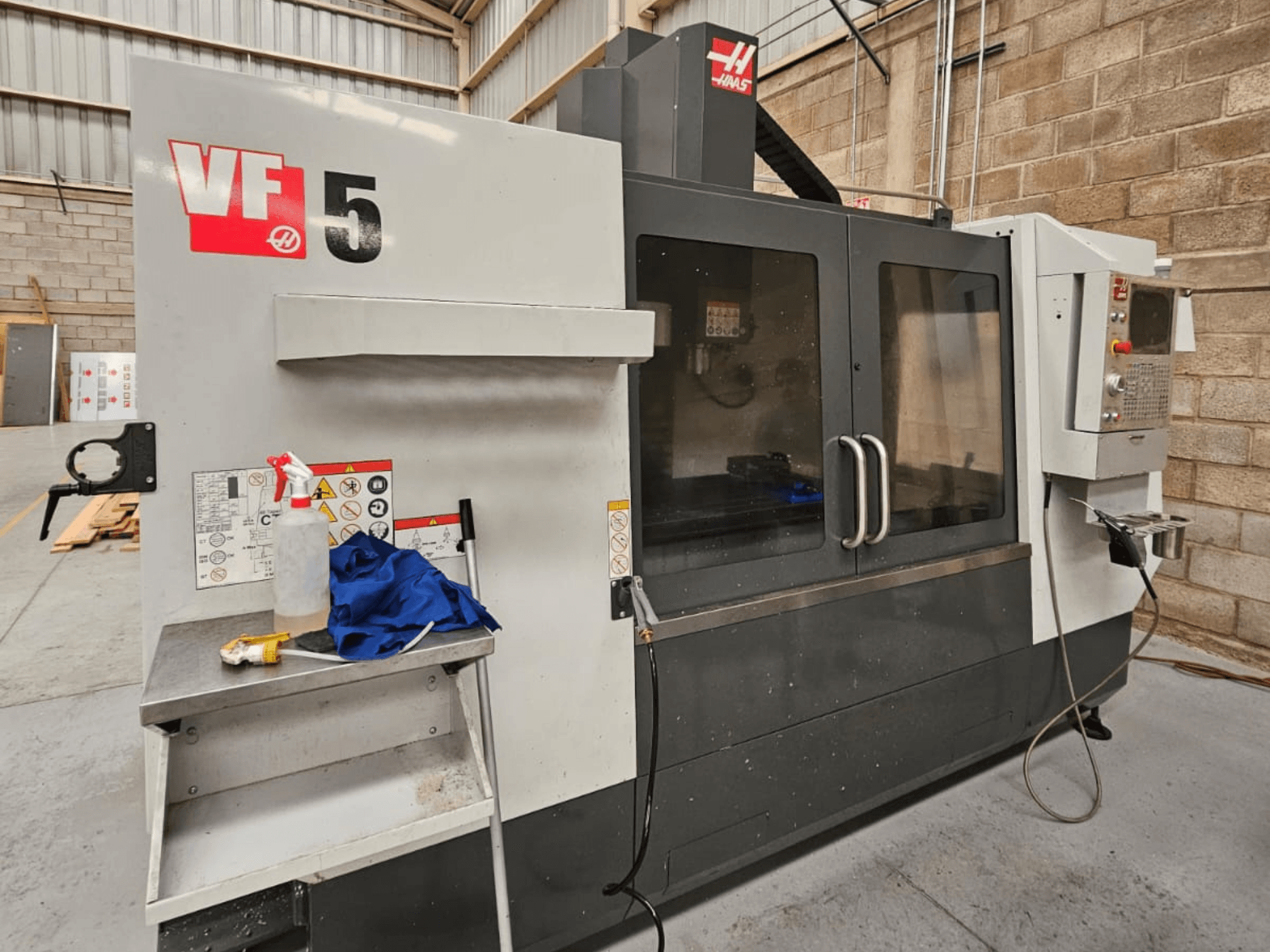 Fresadora CNC Haas VF-5 en vista frontal, con panel de control, botella de refrigerante, paño azul e instrucciones de seguridad.