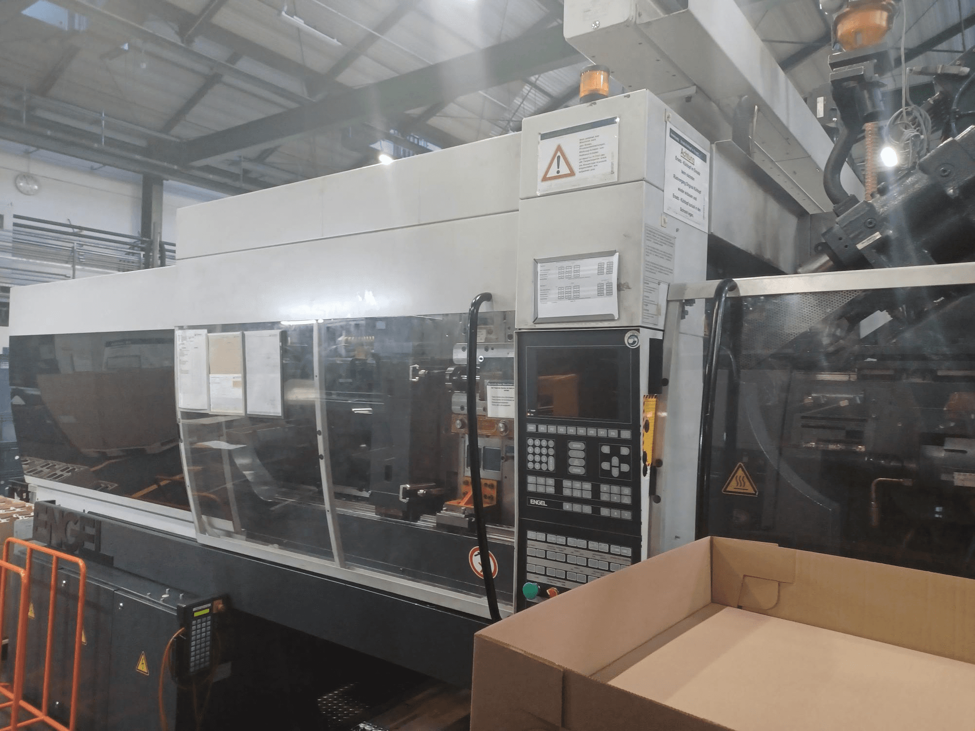 Engel injection molding machine, front view, with control panel, safety labels, and an empty box positioned nearby.