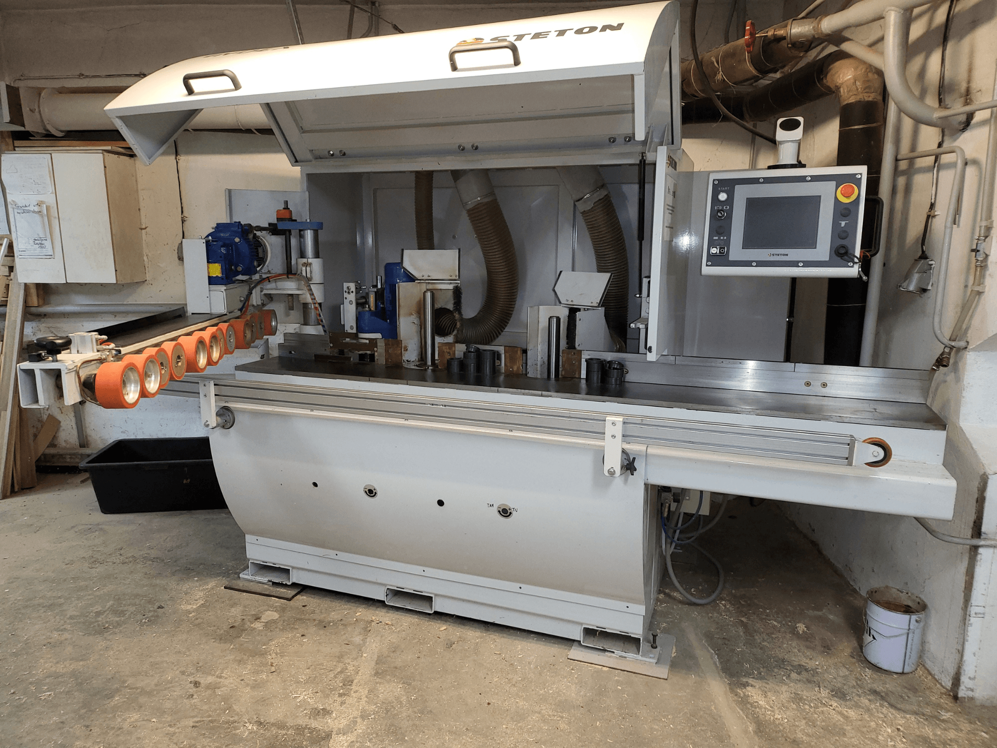 Vista frontal de una máquina para trabajar la madera Stetton con un panel de control, tubos alimentadores y varias piezas de madera sobre la mesa.