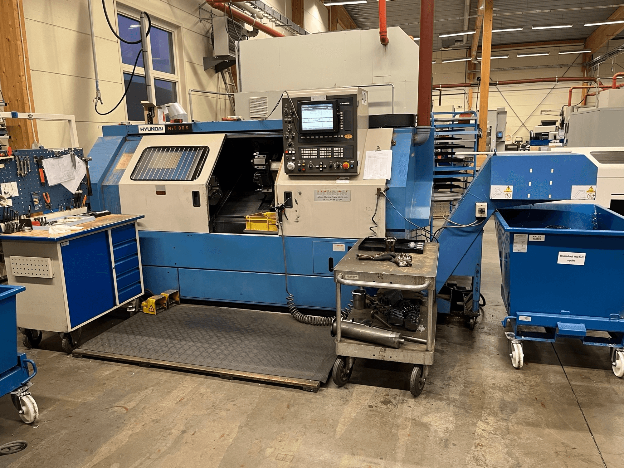 Máquina de torneado horizontal Hyundai HIT 30S, vista frontal, con panel de control y herramientas sobre un carro cercano en un taller.