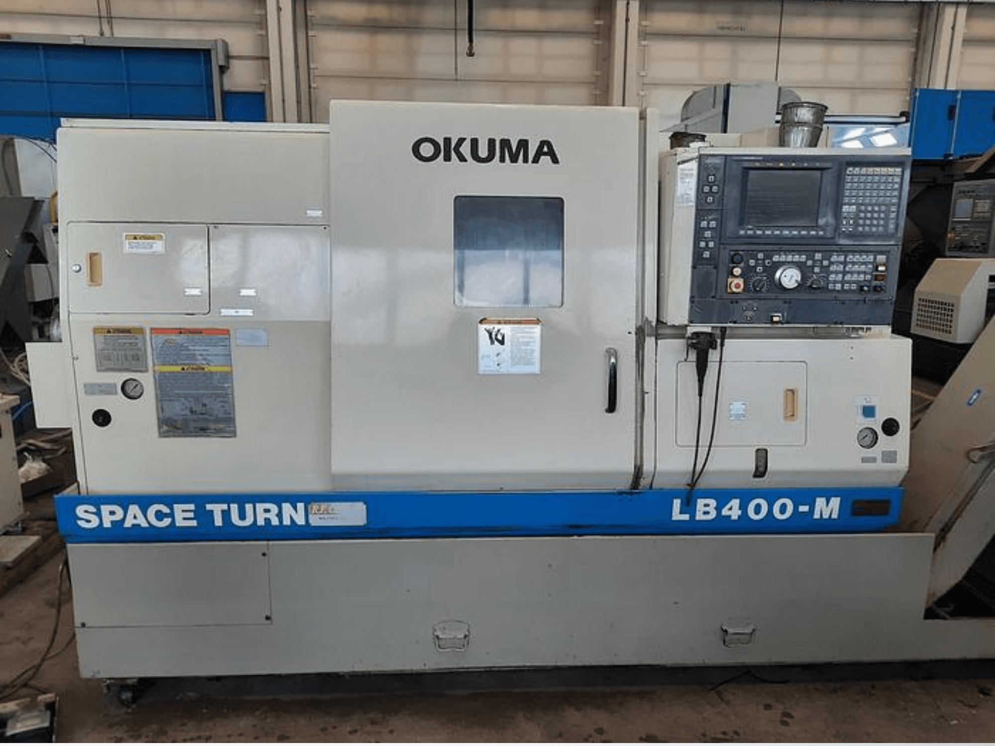 Máquina de torno CNC Okuma LB400-M, vista frontal con panel de control, etiquetas de advertencia y marca azul "SPACE TURN" visible.