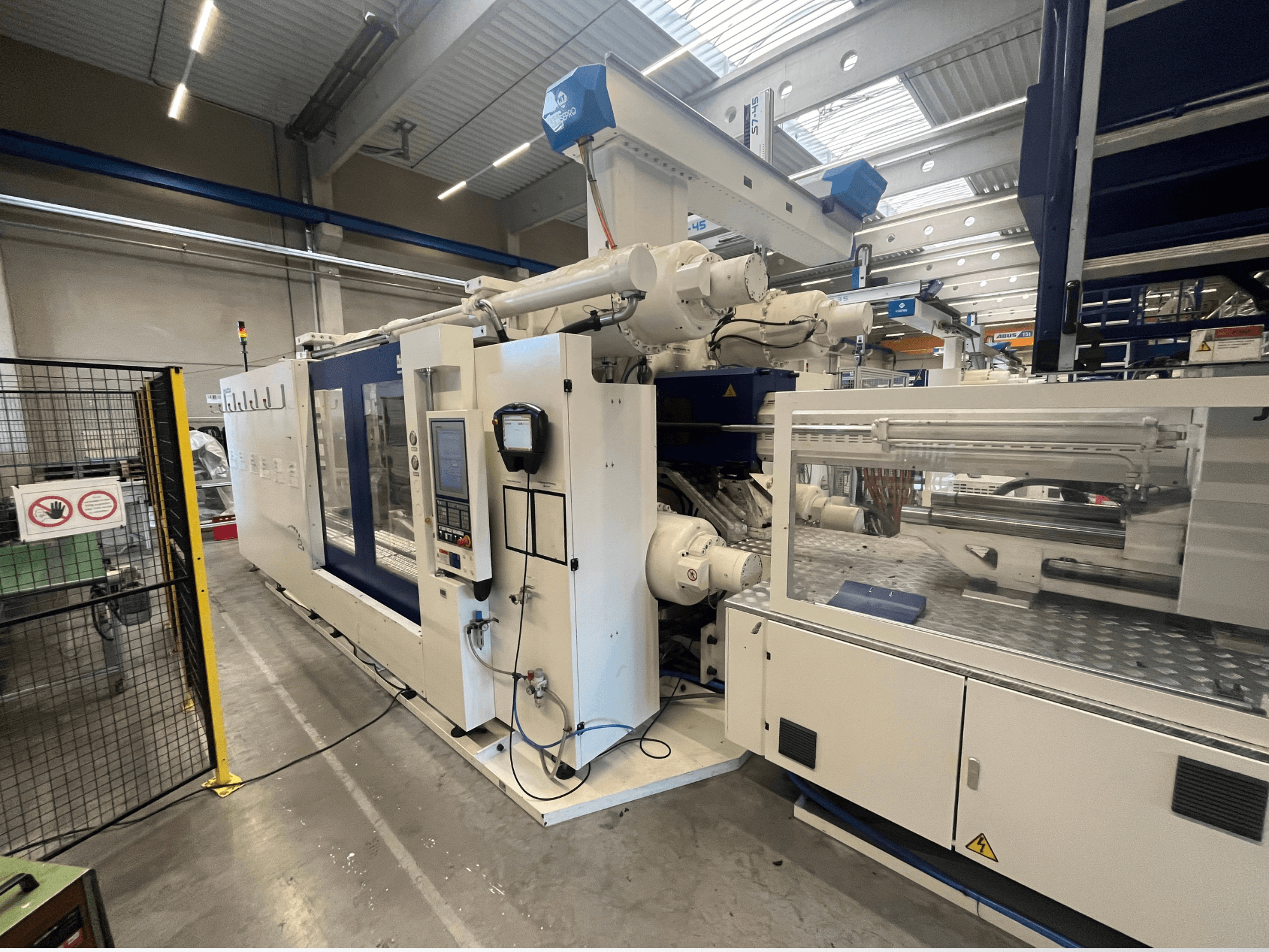 Máquina de moldeo por inyección en blanco y azul, vista frontal, con panel de control, brazo robótico y señalización de seguridad.