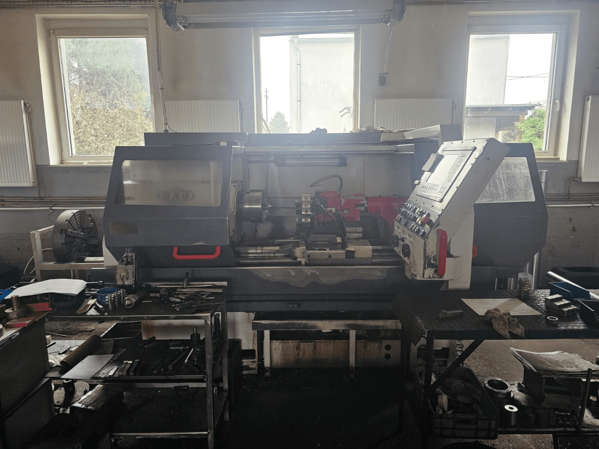 Máquina de torno en un taller, vista frontal, con panel de control y herramientas sobre mesas, rodeada de ventanas y espacio de trabajo desordenado.