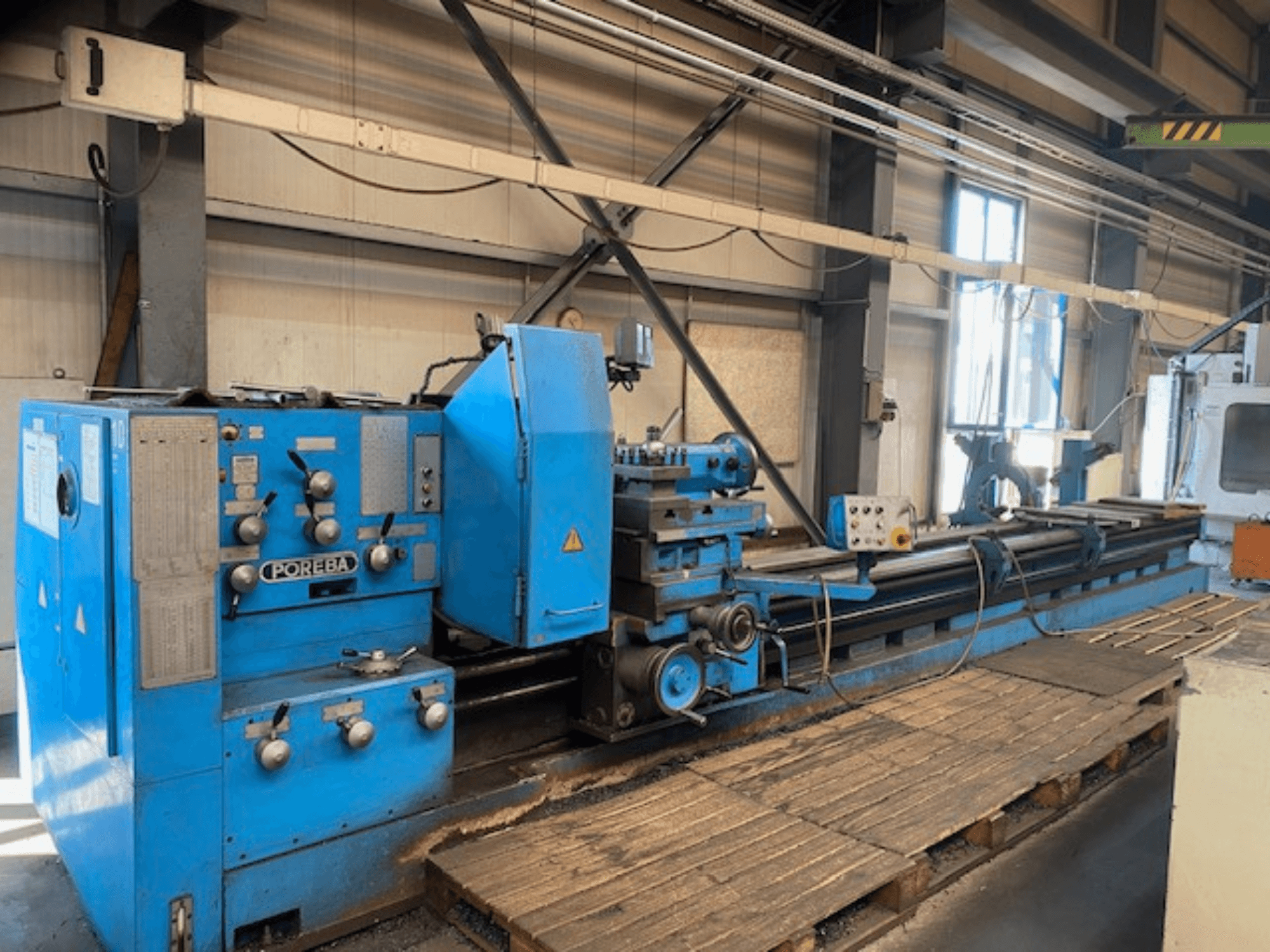 Vista frontal de una máquina de torno POREBA azul, con mandos de control, un soporte para herramientas y palés de madera debajo.