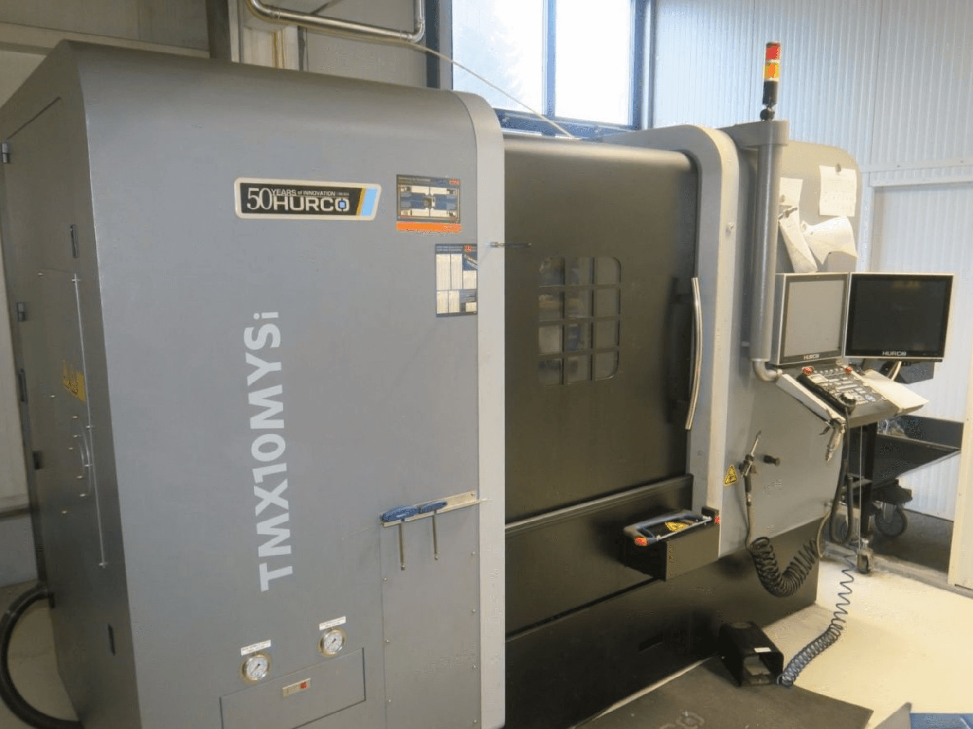 Máquina CNC Hurco TMX10MYSi en vista lateral; presenta panel de control, luces de emergencia e indicadores visualizados.