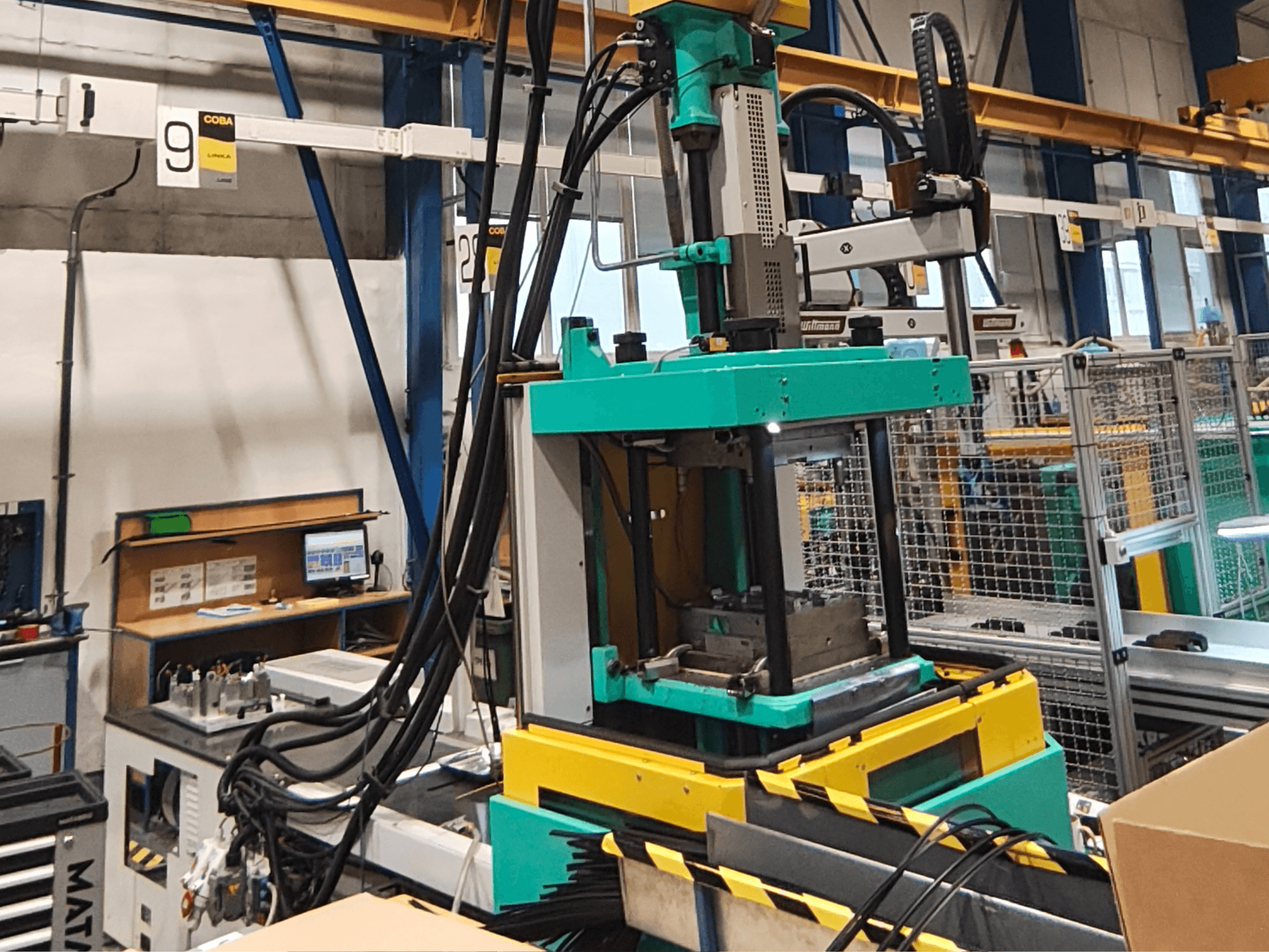 Green and yellow hydraulic press machine in a factory, front view, with visible control panel and cables on the side.