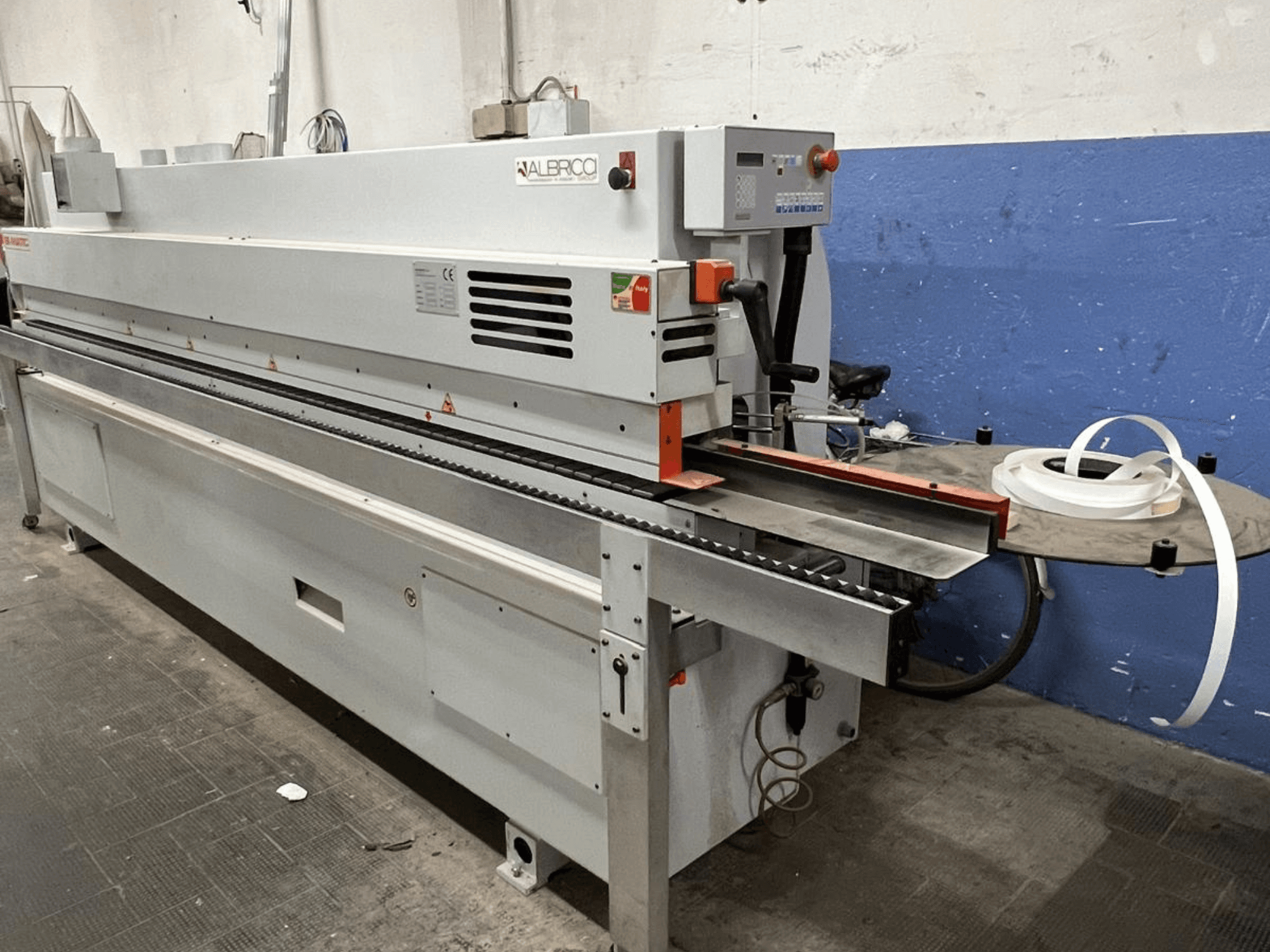 Encoladora de cantos ALBRICO en vista frontal, con una larga cinta transportadora y un rollo de cinta adhesiva acoplado.