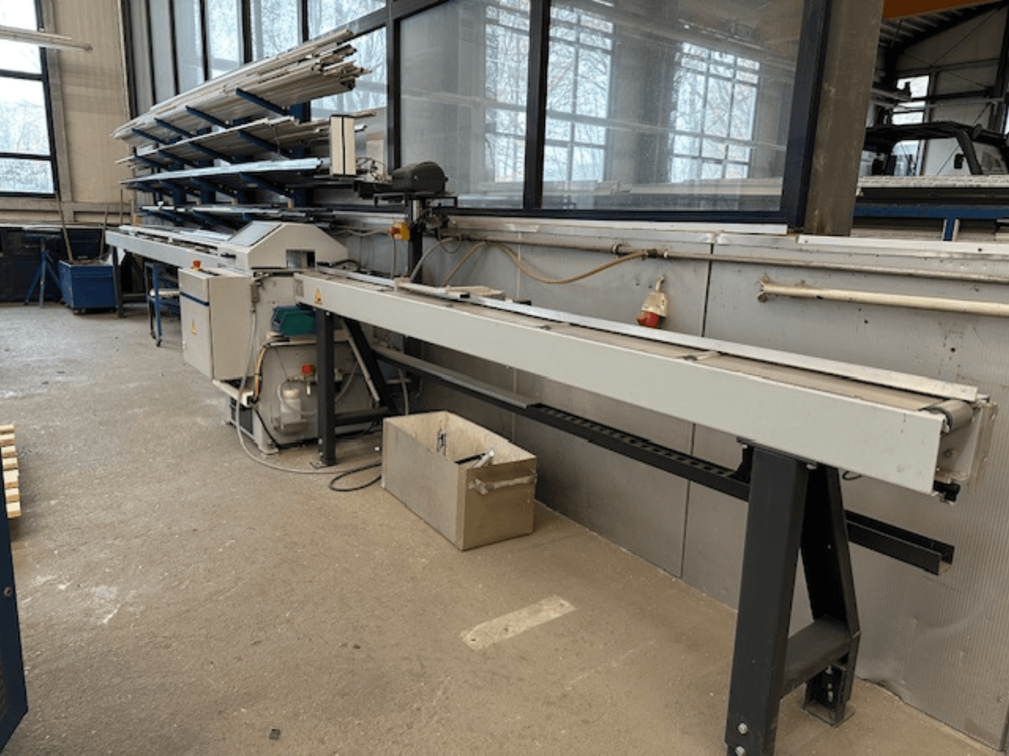 Sierra circular para metal SCHÜCO PGS en un taller, vista lateral con una bancada de corte ajustable y un estante de perfiles metálicos encima.