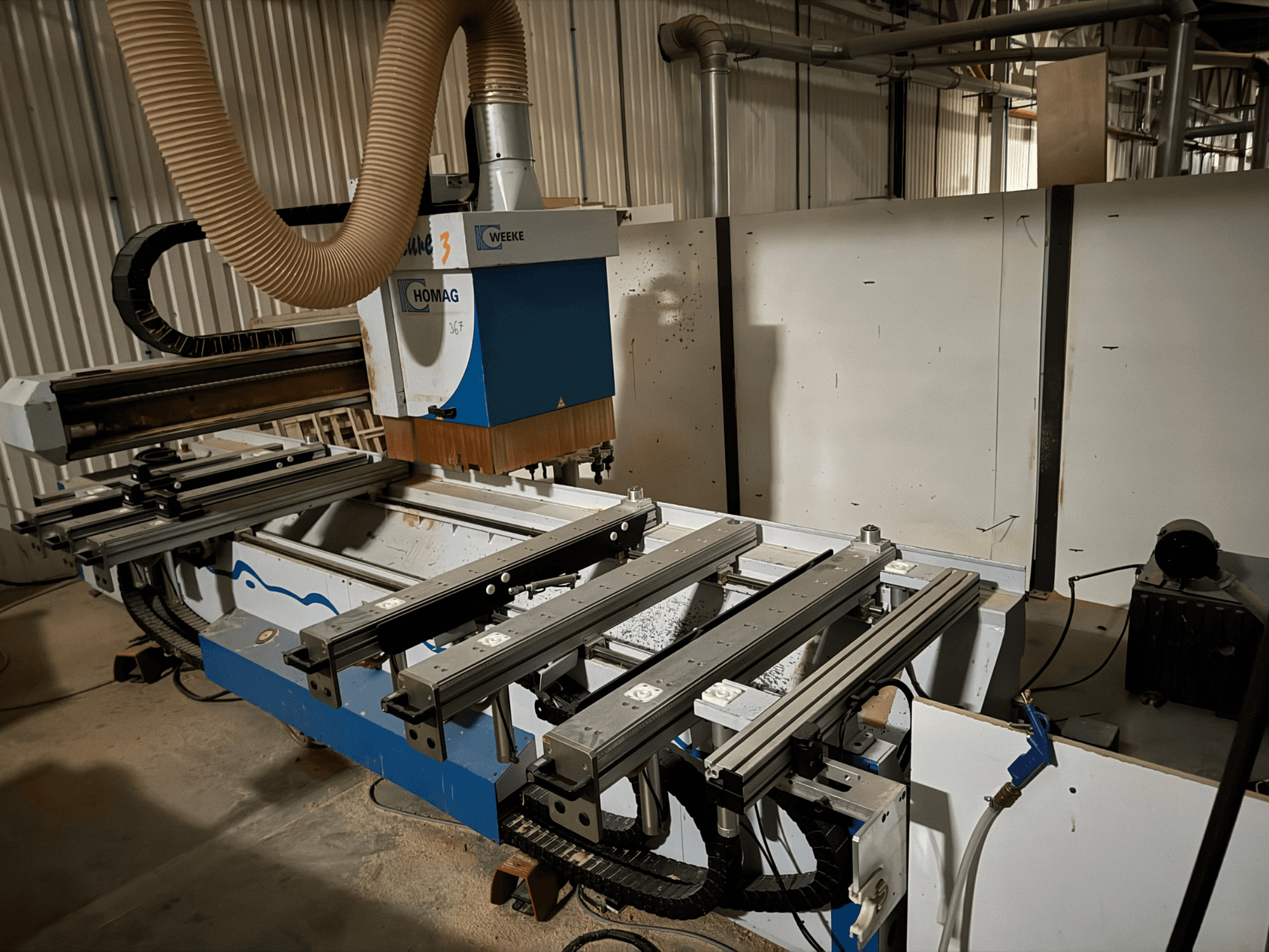 Máquina CNC HOMAG con un diseño en azul y blanco, vista lateral, cuenta con raíles deslizantes, un sistema de extracción de polvo y herramientas montadas.