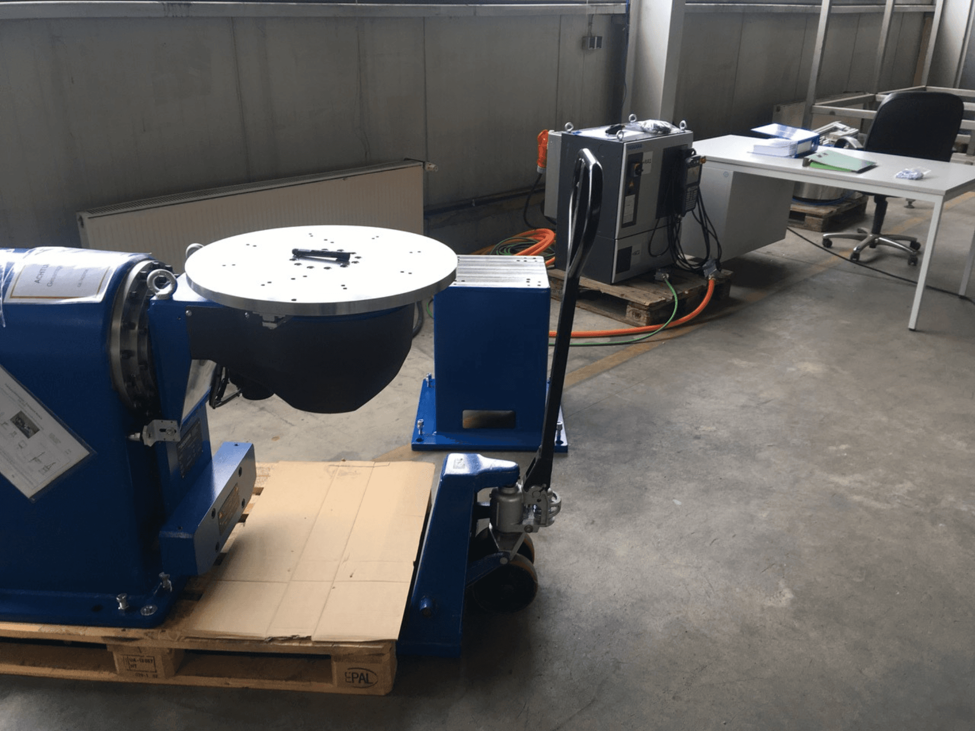 Equipo industrial azul con base circular y panel de control, visto de frente, sobre un palé de madera en un entorno de oficina.