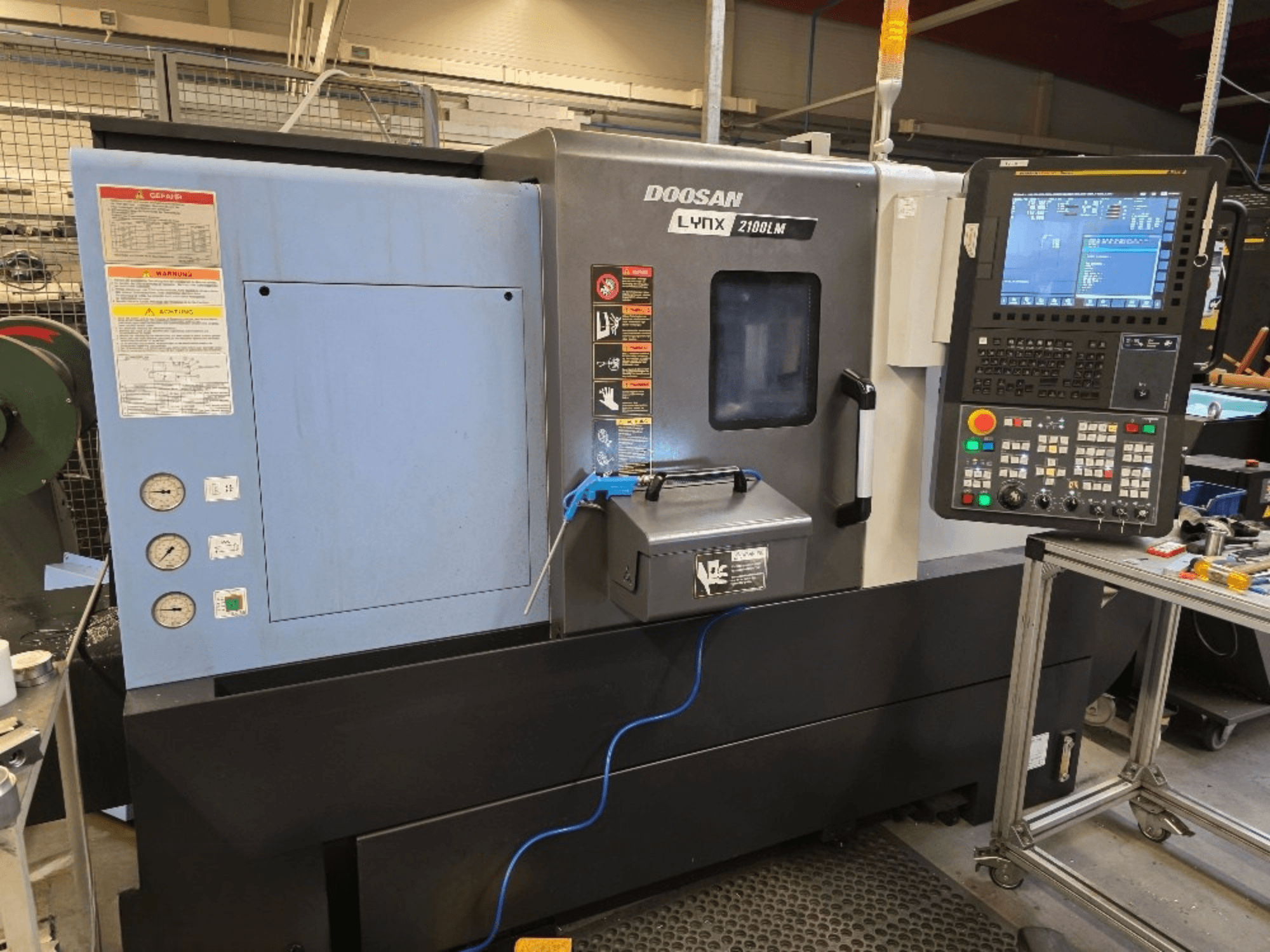 Torno CNC Doosan Lynx 2100LM, vista frontal, con panel de control, manómetros y advertencias de seguridad.