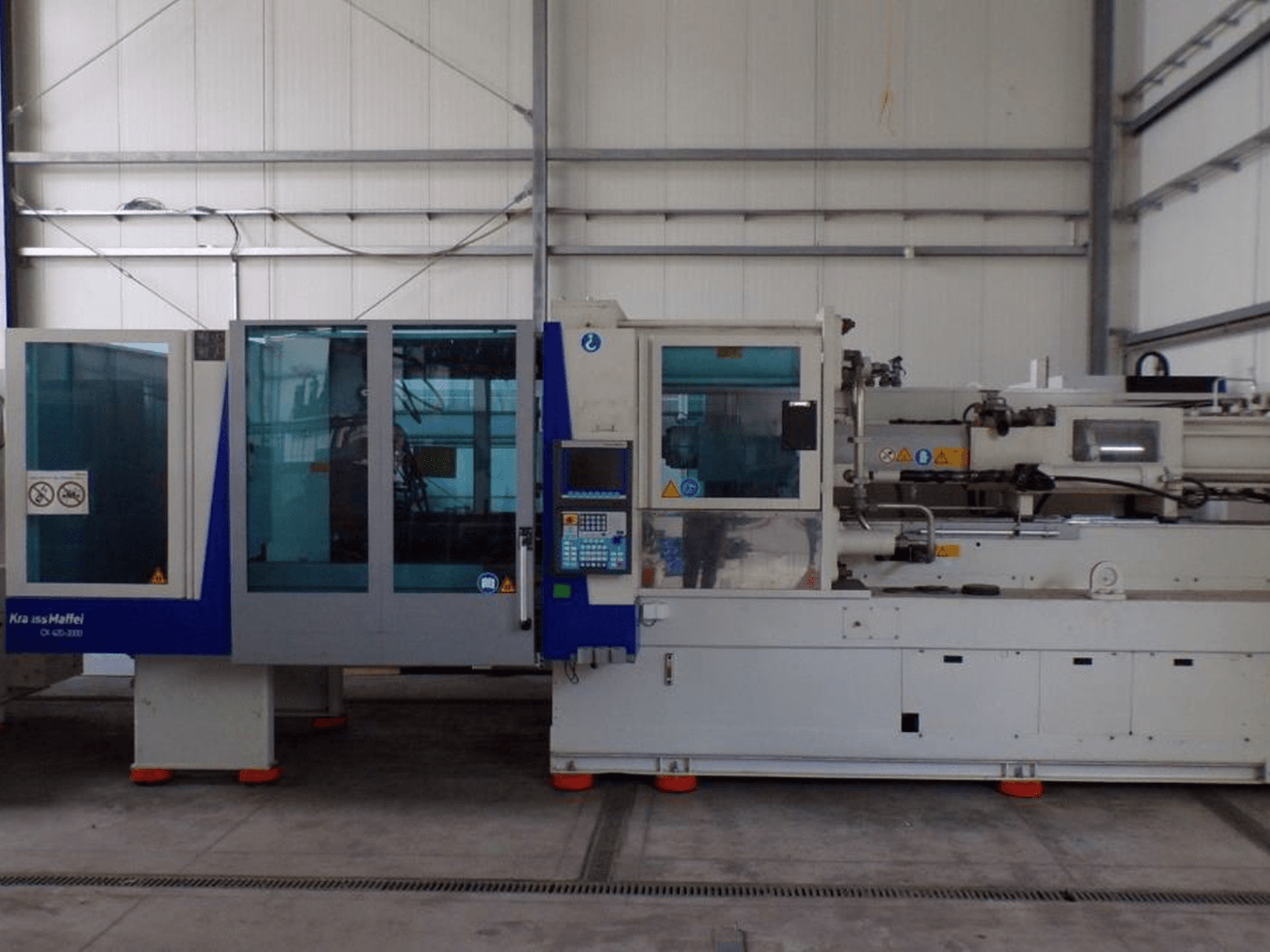 Máquina de moldeo por inyección KraussMaffei, vista lateral, con panel de control e indicadores de seguridad, visible en un entorno de fábrica.