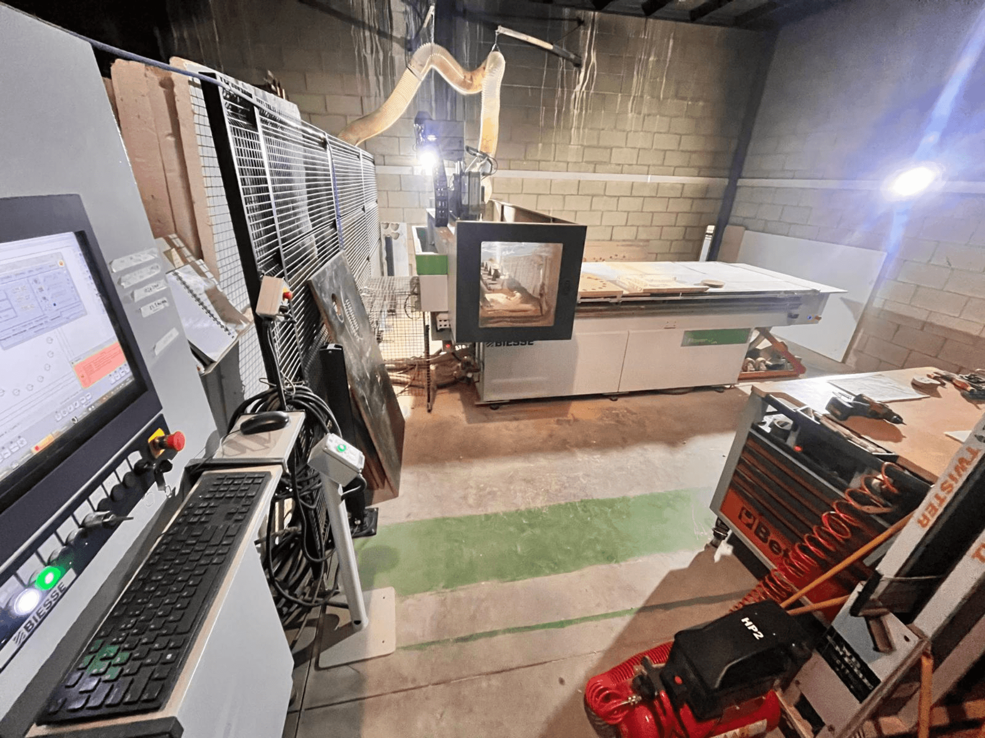 Máquina CNC Biesse en un taller, vista lateral que muestra el panel de control, la extracción de polvo, las herramientas y diversos materiales sobre la mesa.