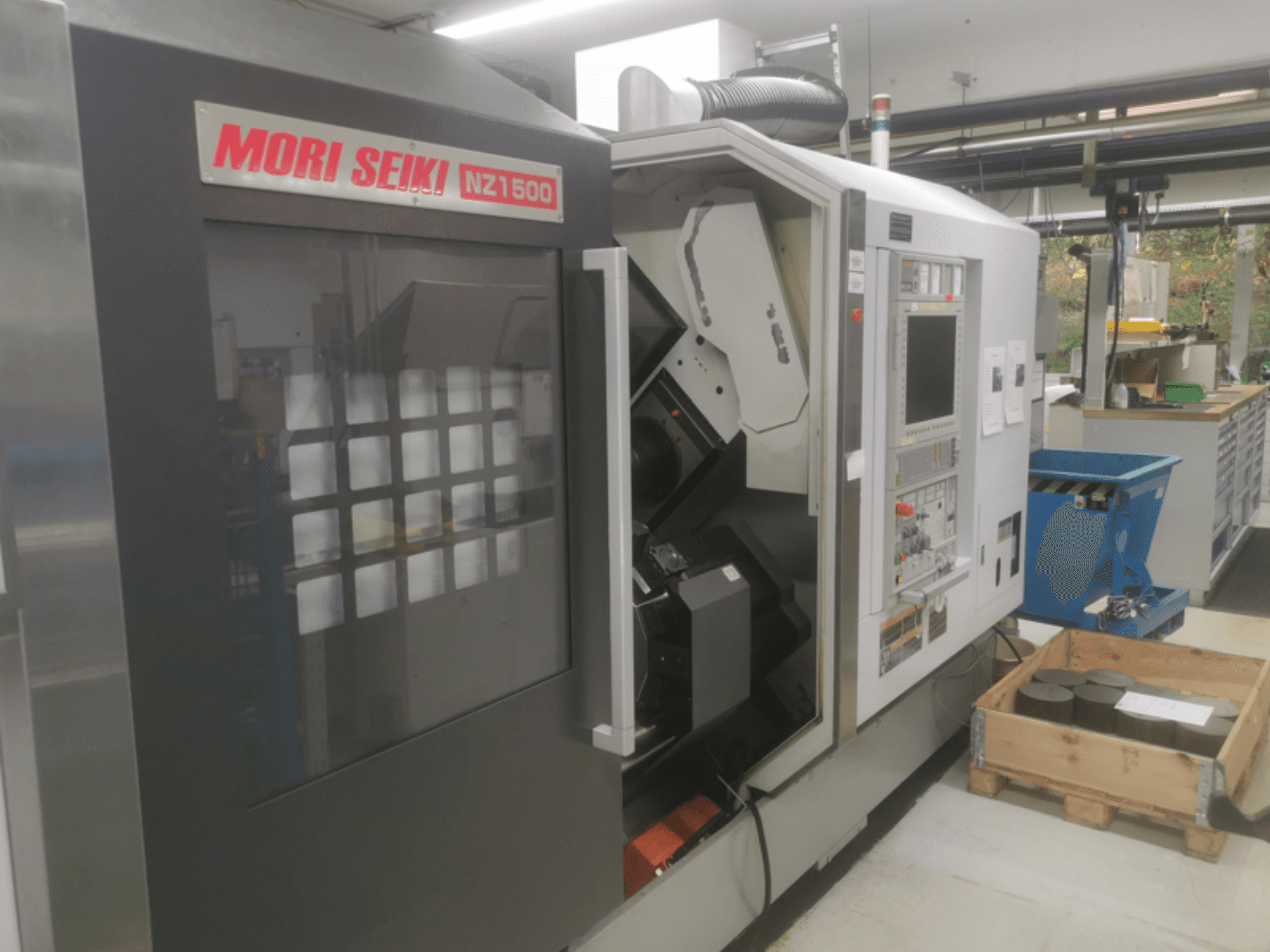 Máquina CNC MORI SEIKI NZ1500 ligeramente inclinada, mostrando el panel de control y el espacio de trabajo con piezas metálicas sobre un palé de madera.