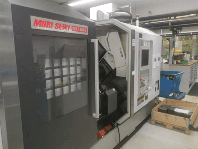 Máquina CNC MORI SEIKI NZ1500 ligeramente inclinada, mostrando el panel de control y el espacio de trabajo con piezas metálicas sobre un palé de madera.