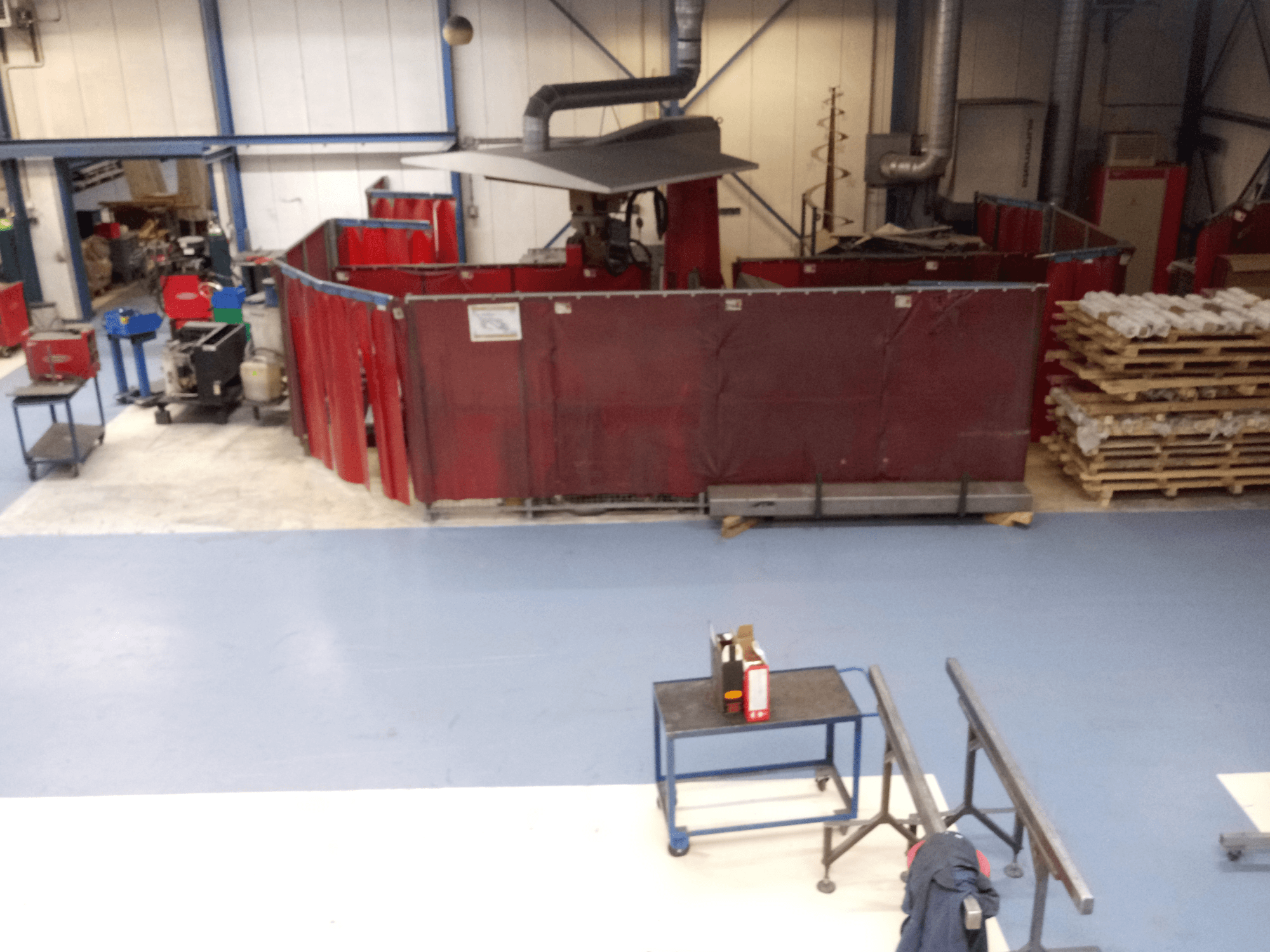 Industrial workspace featuring red welding curtains, machines, and pallets; overhead view showcasing layout and equipment.