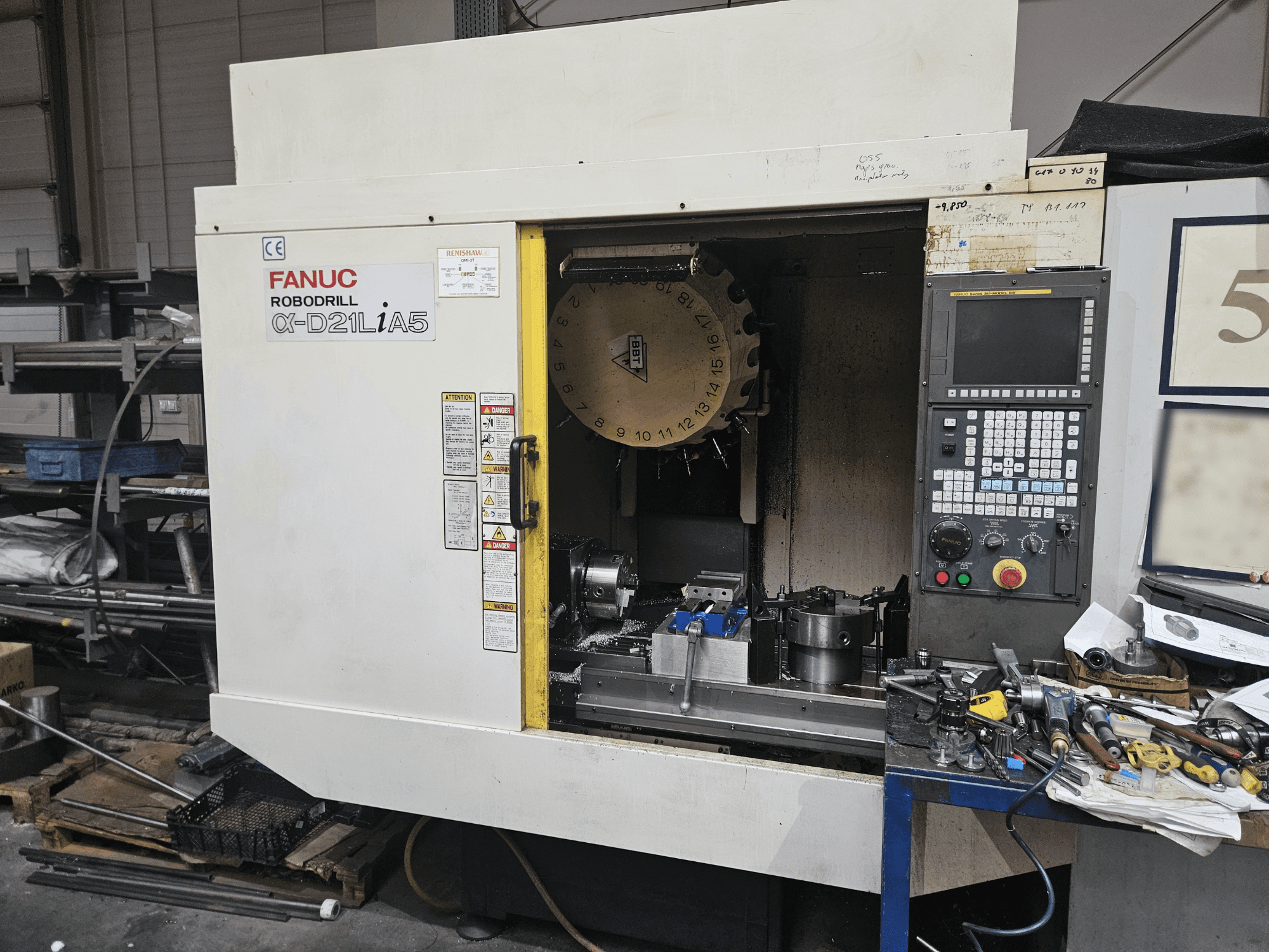 FANUC Robodrill α-D21LiA5, vista frontal que muestra el portaherramientas y el panel de control, rodeado de herramientas y equipos en un taller.